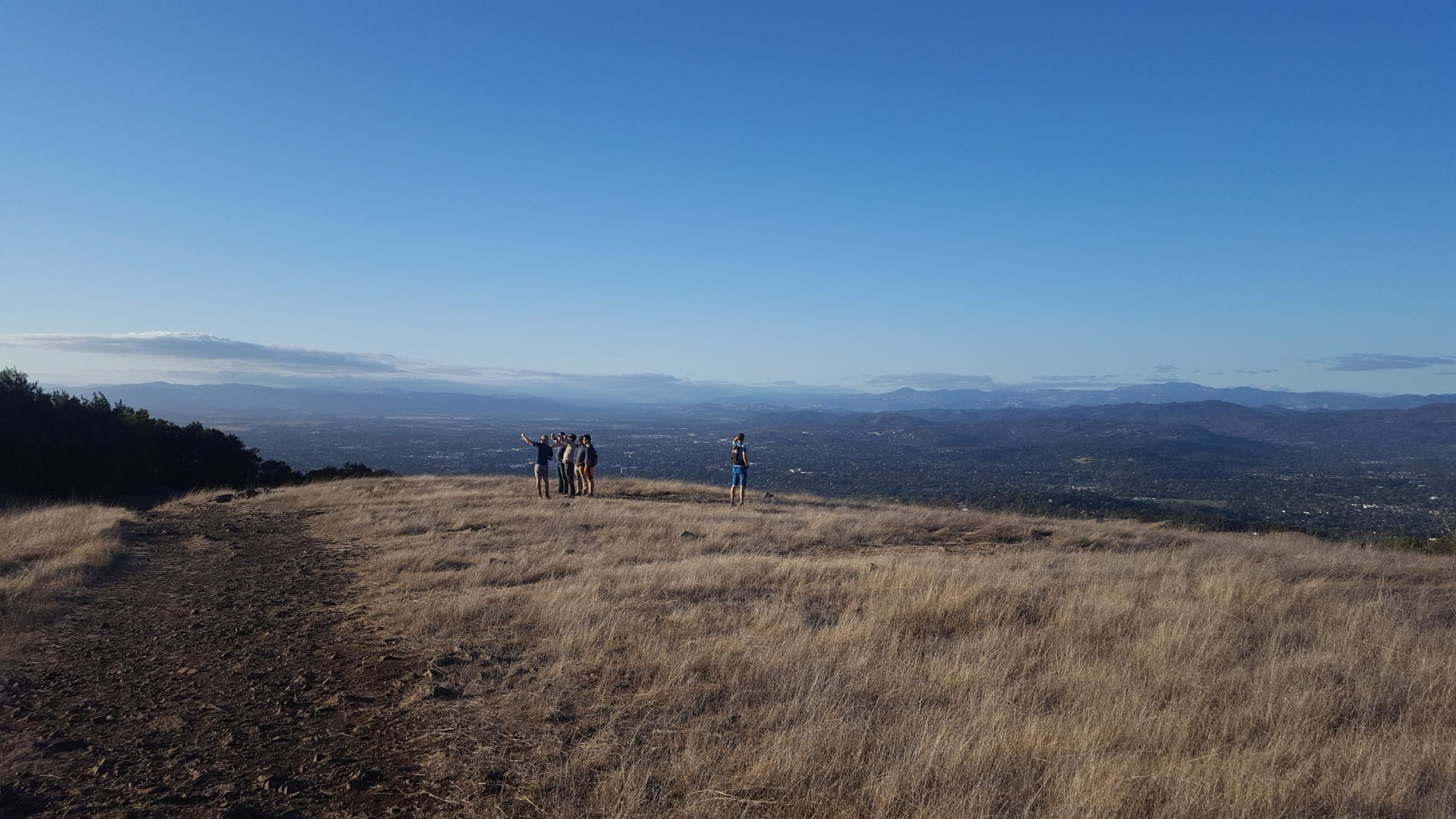 A group of people standing on a hillside, enjoying a panoramic view of the valley below, with distant mountains visible under a clear blue sky. The terrain is dry with tufts of grass, and the trail is rocky, indicating a hiking area. Taylor Mountain mountain bike trail.