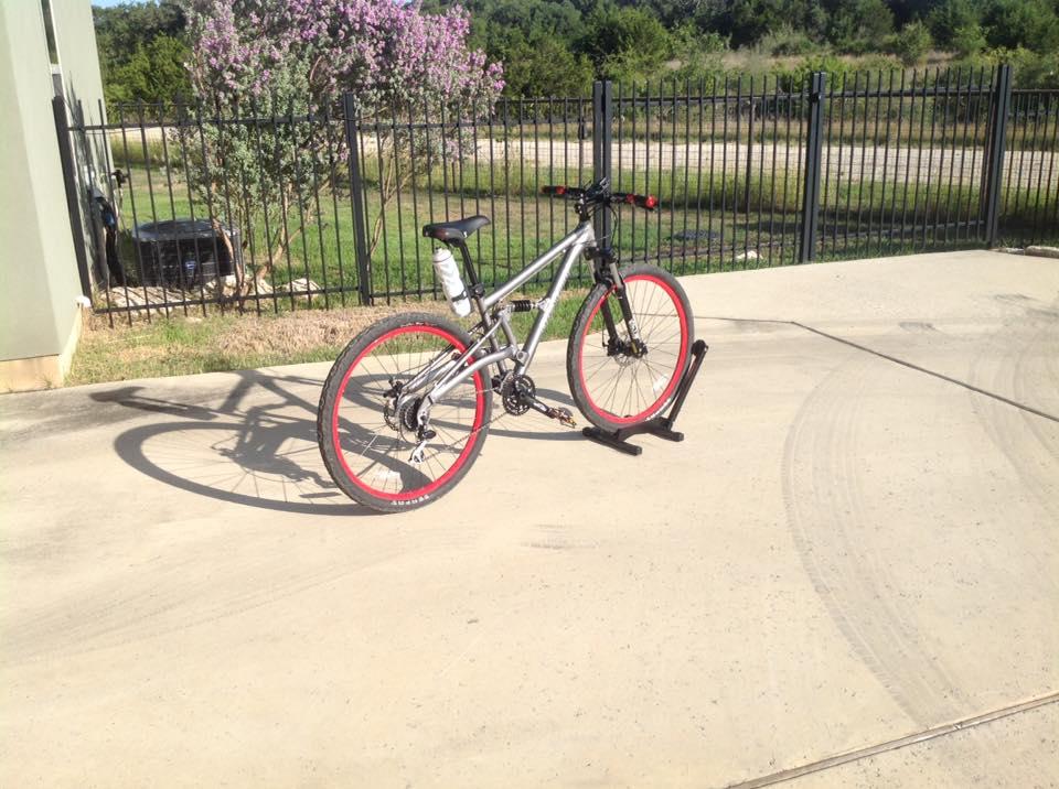 Gravity FSX 29One: A silver mountain bike with red rims is parked on a concrete surface. It stands on a black bike stand, with a background of green grass and a black fence. There are trees in the distance, and the scene is well-lit by sunlight.