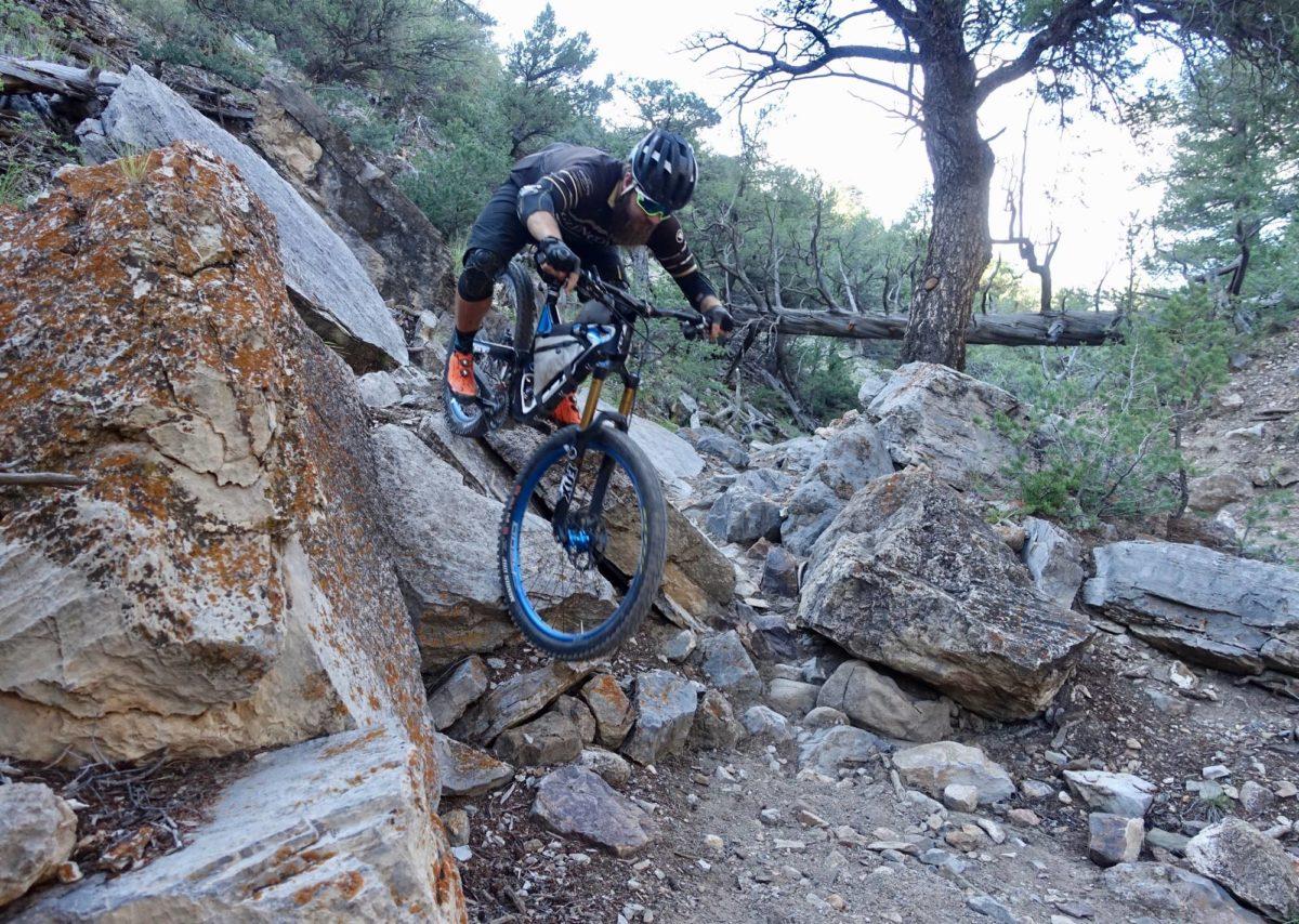 Michelin Wild AM: A mountain biker navigating a rocky trail, jumping off a boulder with trees and rugged terrain in the background. The biker is wearing protective gear and has a focused expression as they tackle the challenging path.