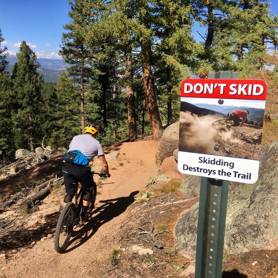 A mountain biker in a yellow helmet rides along a dirt trail surrounded by tall trees. In the foreground, a sign reads "DON'T SKID" with the message "Skidding Destroys the Trail" along with an image of a biker skidding on a rocky path. Little Scraggy mountain bike trail.