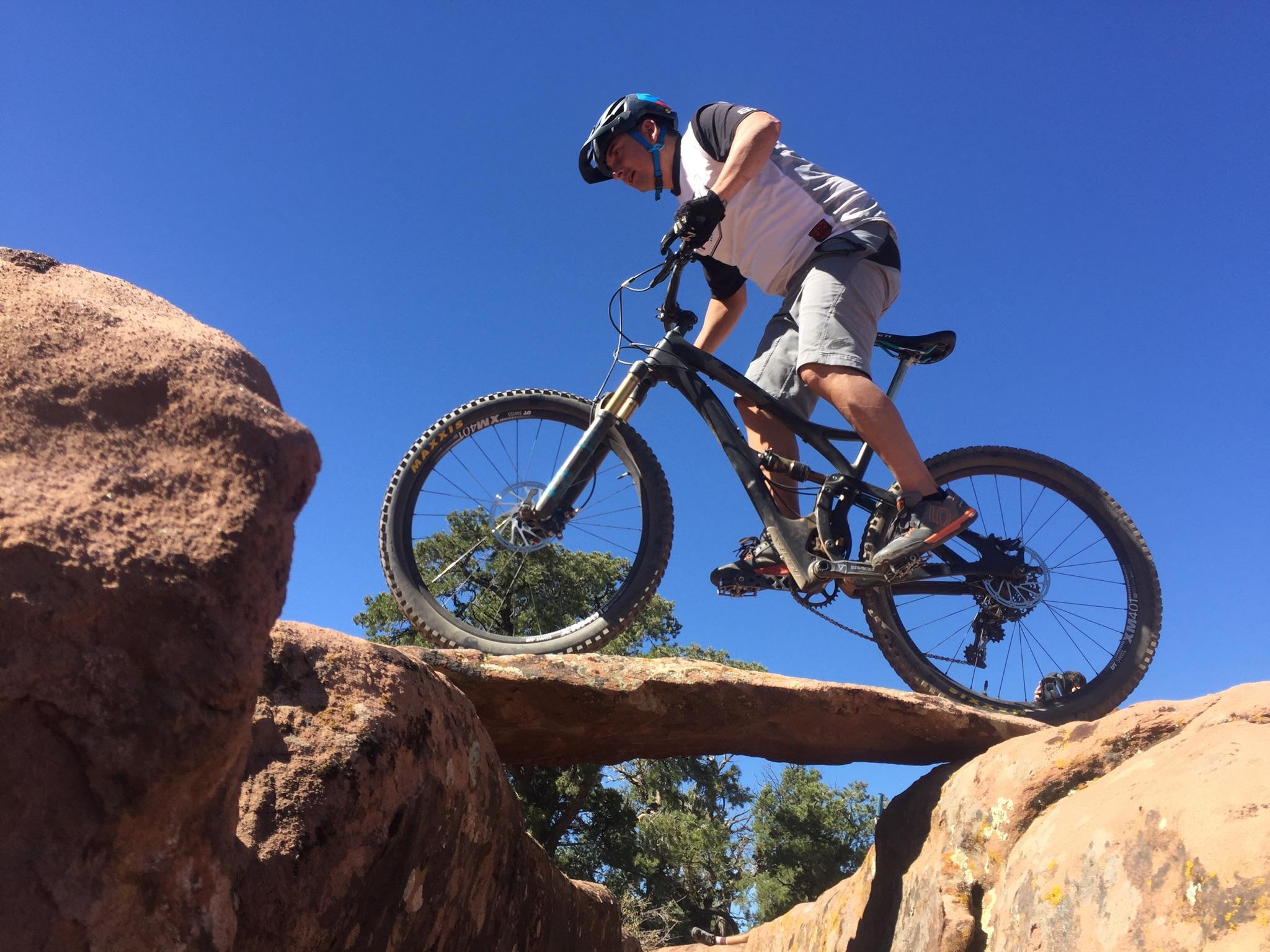 Specialized Stumpjumper FSR comp carbon 650B: A mountain biker skillfully navigating a rocky terrain, balancing on a narrow rock ledge against a clear blue sky. The rider is wearing a helmet and cycling gear, demonstrating expertise in mountain biking. Surrounding the rocks are patches of greenery, adding contrast to the rugged landscape.