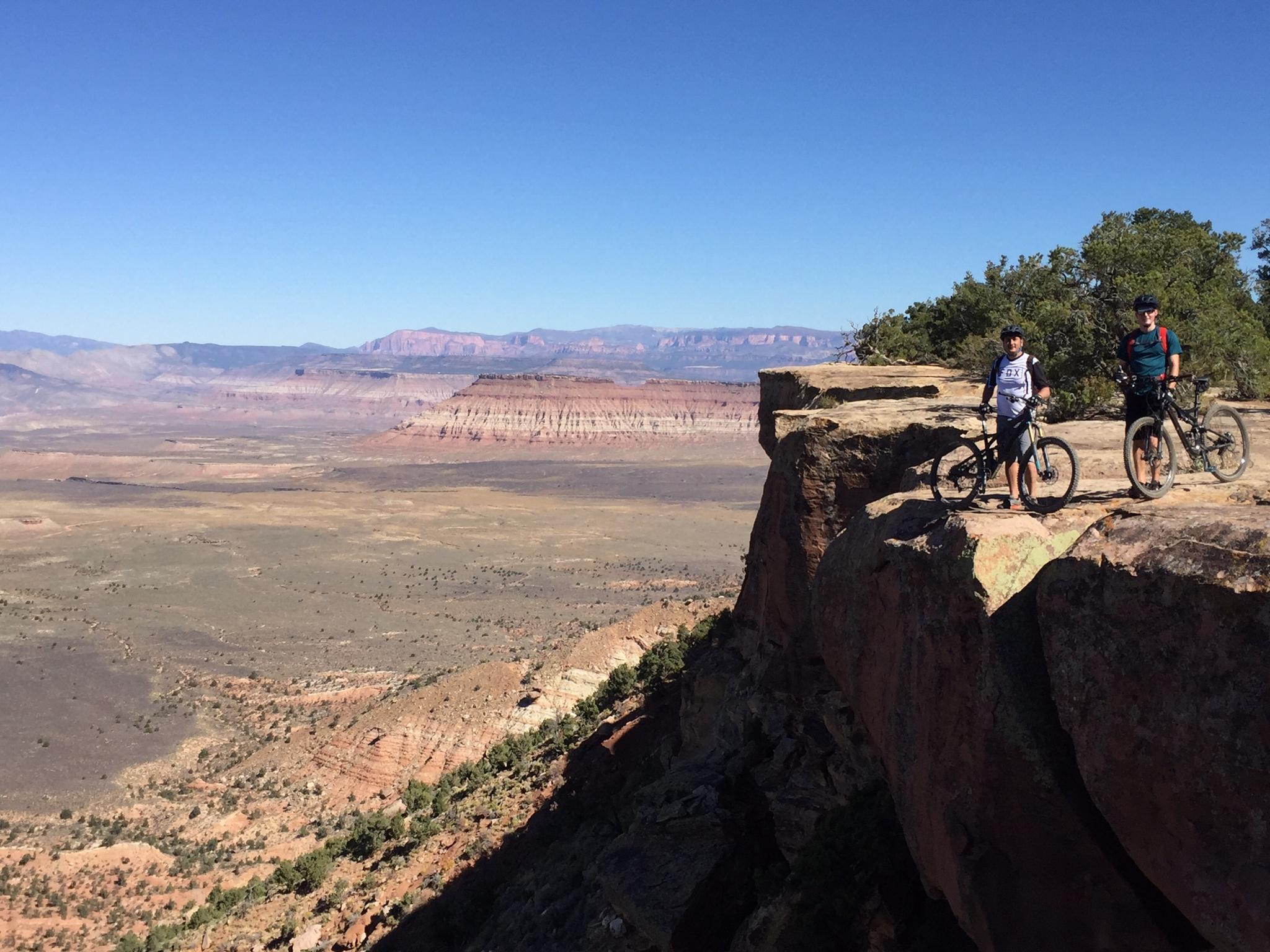 Specialized Stumpjumper FSR comp carbon 650B: Two mountain bikers standing on a rocky ledge overlooking a vast desert landscape. The scene features rolling hills, colorful rock formations, and a clear blue sky. The bikers are posed with their bikes, enjoying the panoramic view of the terrain.