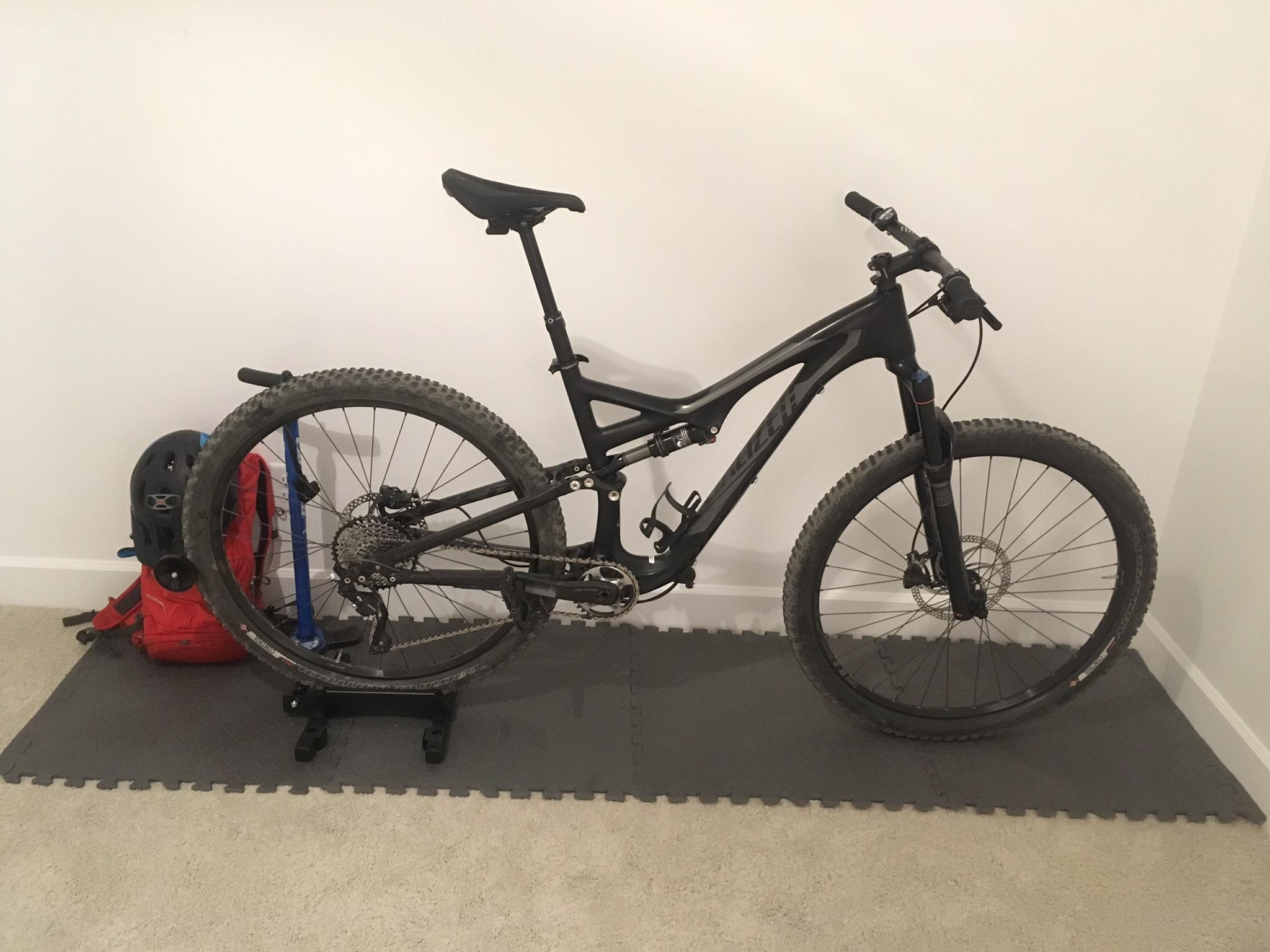Specialized Stumpjumper FSR comp carbon 650B: A black mountain bike parked on a gray exercise mat, next to a red backpack and a blue bike stand. A black helmet is also resting on the backpack, against a plain white wall. The bike features knobby tires and a suspension system, indicating it's designed for rugged terrain.