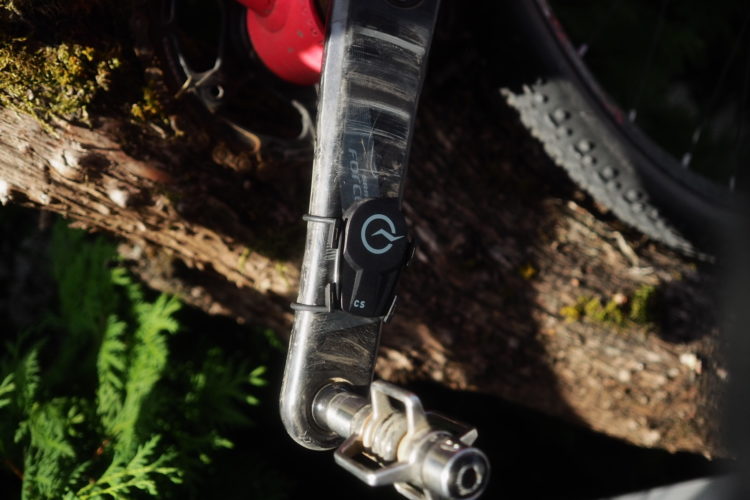 Close-up of a bicycle crank arm with a pedal attached, resting on a moss-covered log. The sunlight highlights the metallic finish of the crank arm and reveals details of the pedal system. Green foliage is visible in the background.