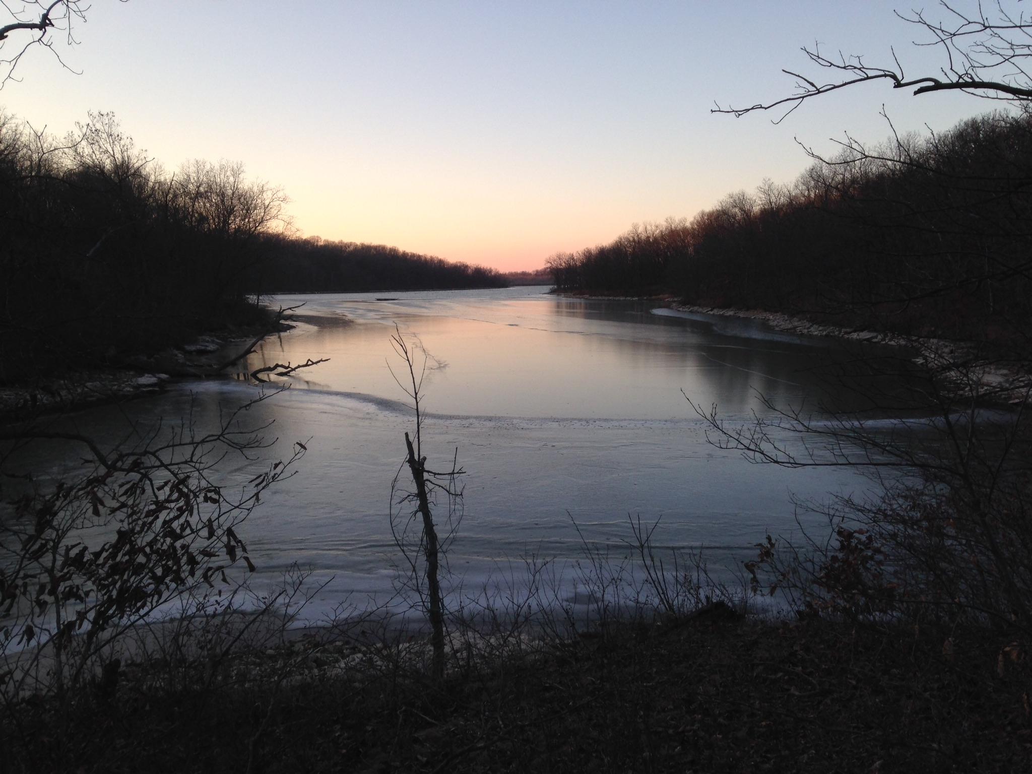 A tranquil river scene at dusk, featuring calm waters reflecting the soft colors of the sky. Bare trees line the banks, with some reaching out over the water. Ice can be seen along the edges of the river, indicating a cool temperature. The fading light creates a serene atmosphere. Thousand Hills mountain bike trail.