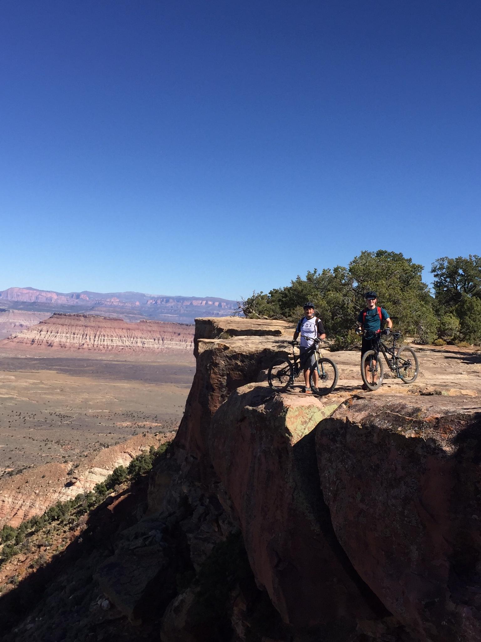 Specialized Stumpjumper FSR comp carbon 650B: Two mountain bikers standing near the edge of a rocky cliff, overlooking a vast landscape with layered hills and a clear blue sky. The riders have their bikes beside them and are dressed in outdoor gear, surrounded by sparse vegetation.