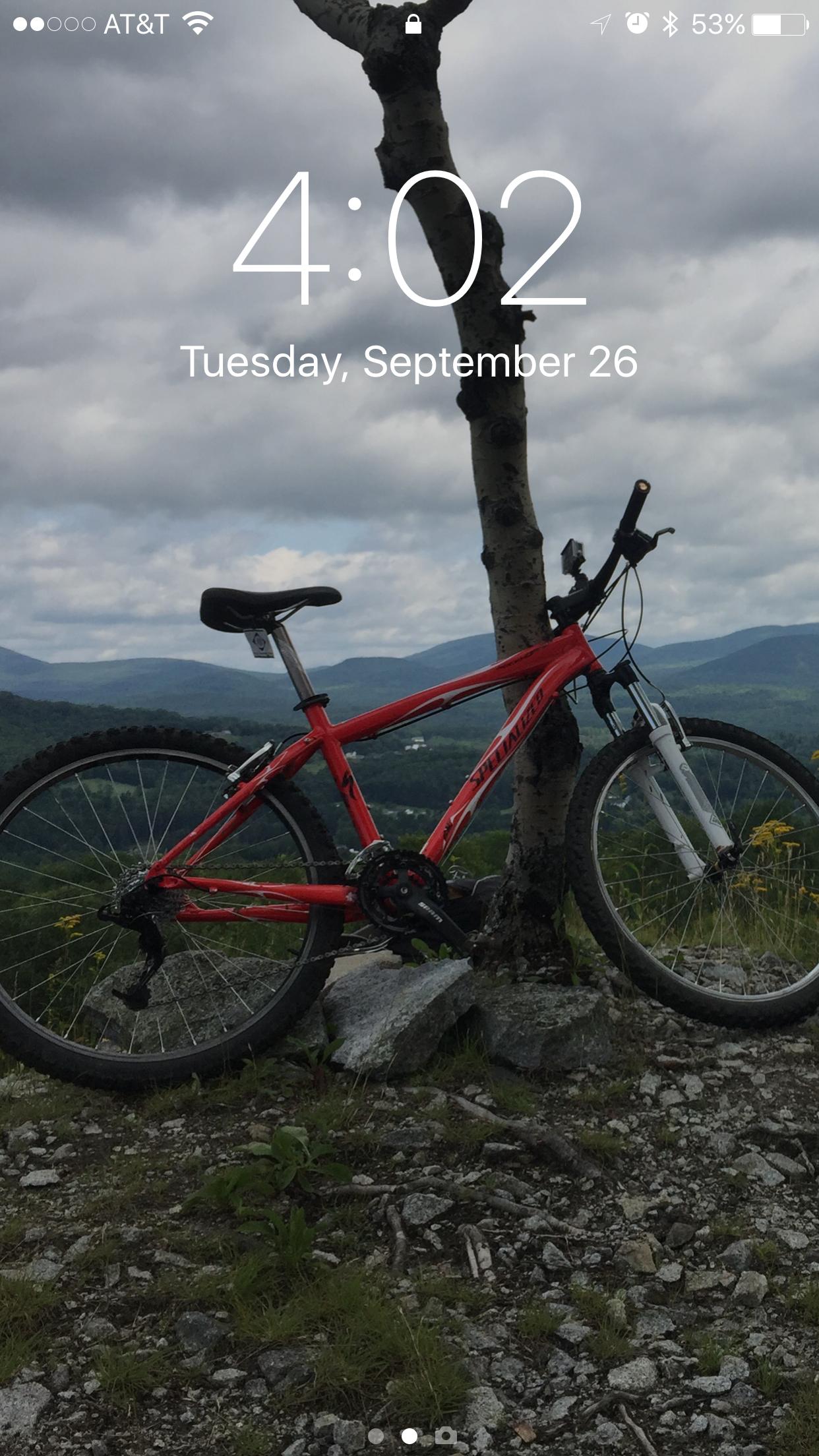 Specialized Rockhopper: A red mountain bike resting against a tree on rocky terrain, with a scenic view of rolling hills and cloudy skies in the background. The time displayed on the device screen is 4:02 PM, and the date is September 26.