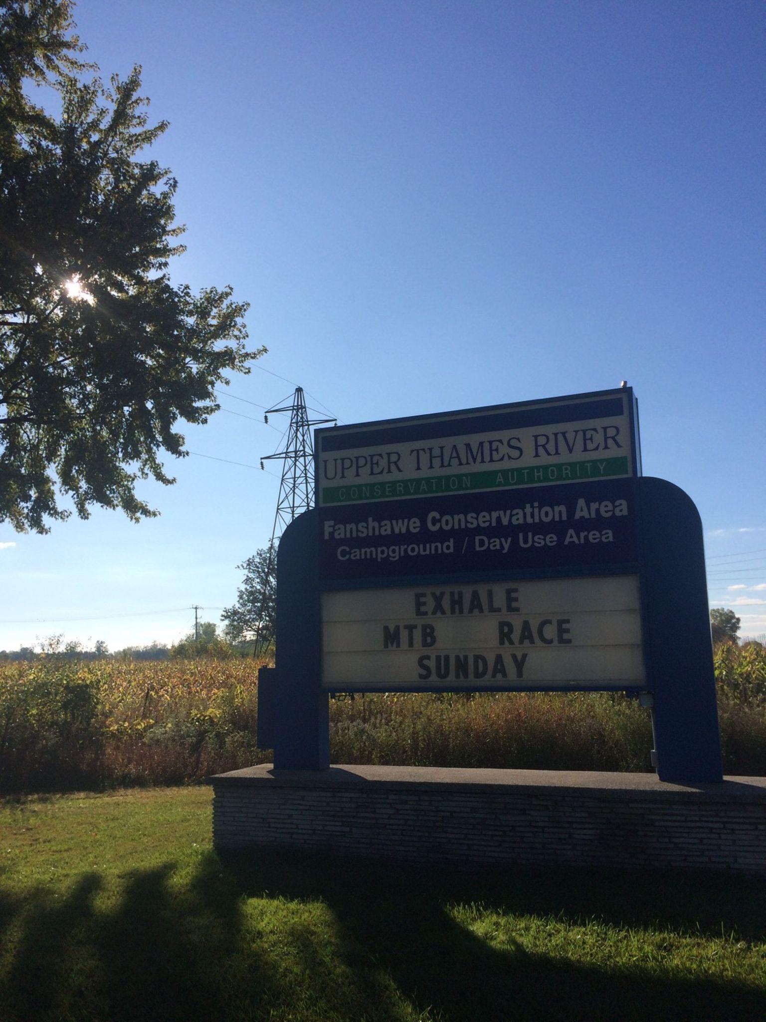 Sign for Fanshawe Conservation Area displaying information about an upcoming mountain bike race, set against a clear blue sky with a power line and trees in the background. Fanshawe Lake mountain bike trail.