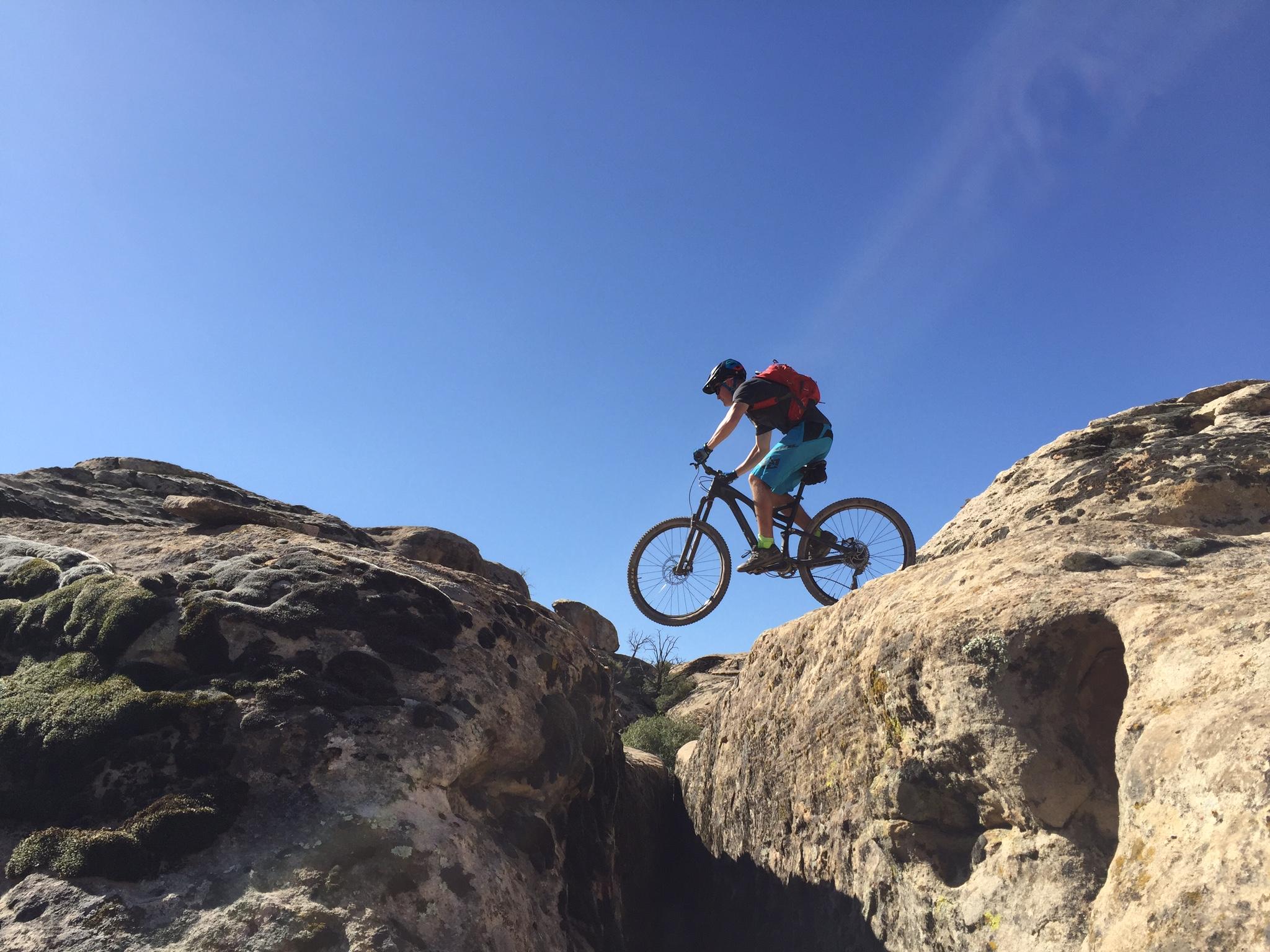 Specialized Stumpjumper FSR comp carbon 650B: A mountain biker jumps over a rocky ledge against a clear blue sky, showcasing a dynamic action shot in an outdoor adventure setting.