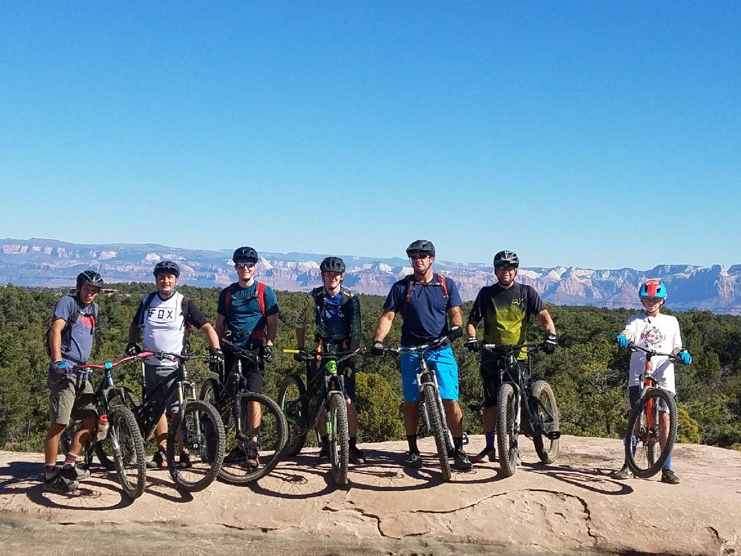 Specialized Stumpjumper FSR comp carbon 650B: A group of seven mountain bikers, including an adult and a child, pose together with their bikes on a rocky outcrop. They are surrounded by a lush green landscape and mountains in the background under a clear blue sky. The cyclists wear helmets and various biking attire, smiling and enjoying the outdoor setting.