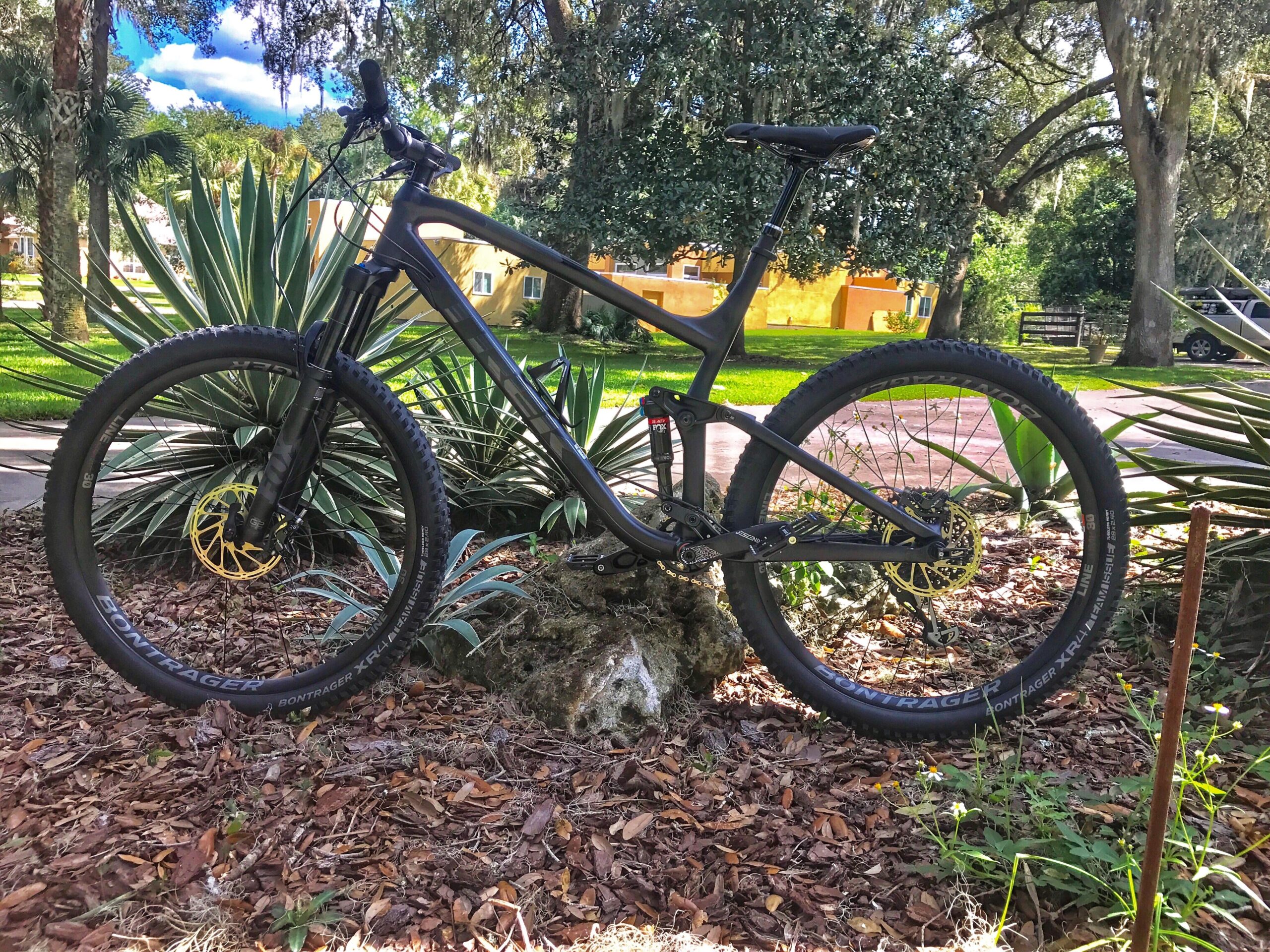 Trek Fuel EX 8: A black mountain bike positioned on a rock, surrounded by lush greenery and tropical plants. In the background, trees and an out-of-focus building with yellow accents are visible under a partly cloudy sky.