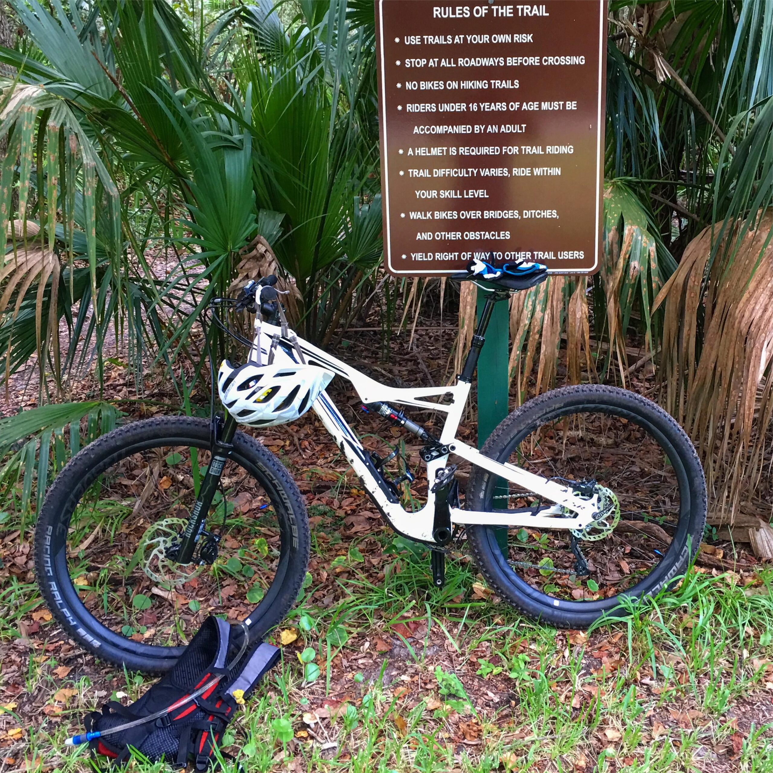 Specialized Stumpjumper FSR comp carbon 650B: A mountain bike leaning against a trail sign surrounded by lush greenery. The sign states rules for trail use, including safety guidelines and restrictions for riders. A helmet rests on the handlebars, and the bike's tires are fitted for rugged terrain.