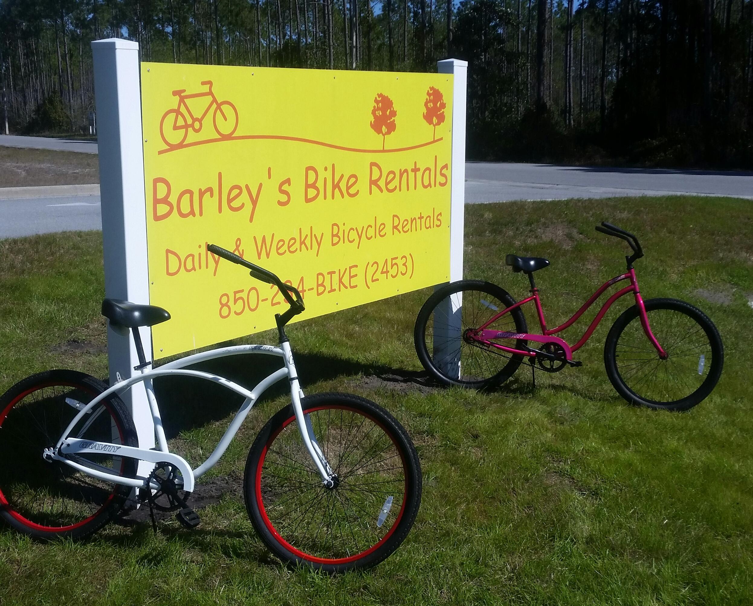 A bright yellow sign for Barley's Bike Rentals displays the business name and rental information, with two bicycles parked in front: one white with red wheels and one pink. The setting features green grass and a backdrop of tall trees, indicating a scenic outdoor location.