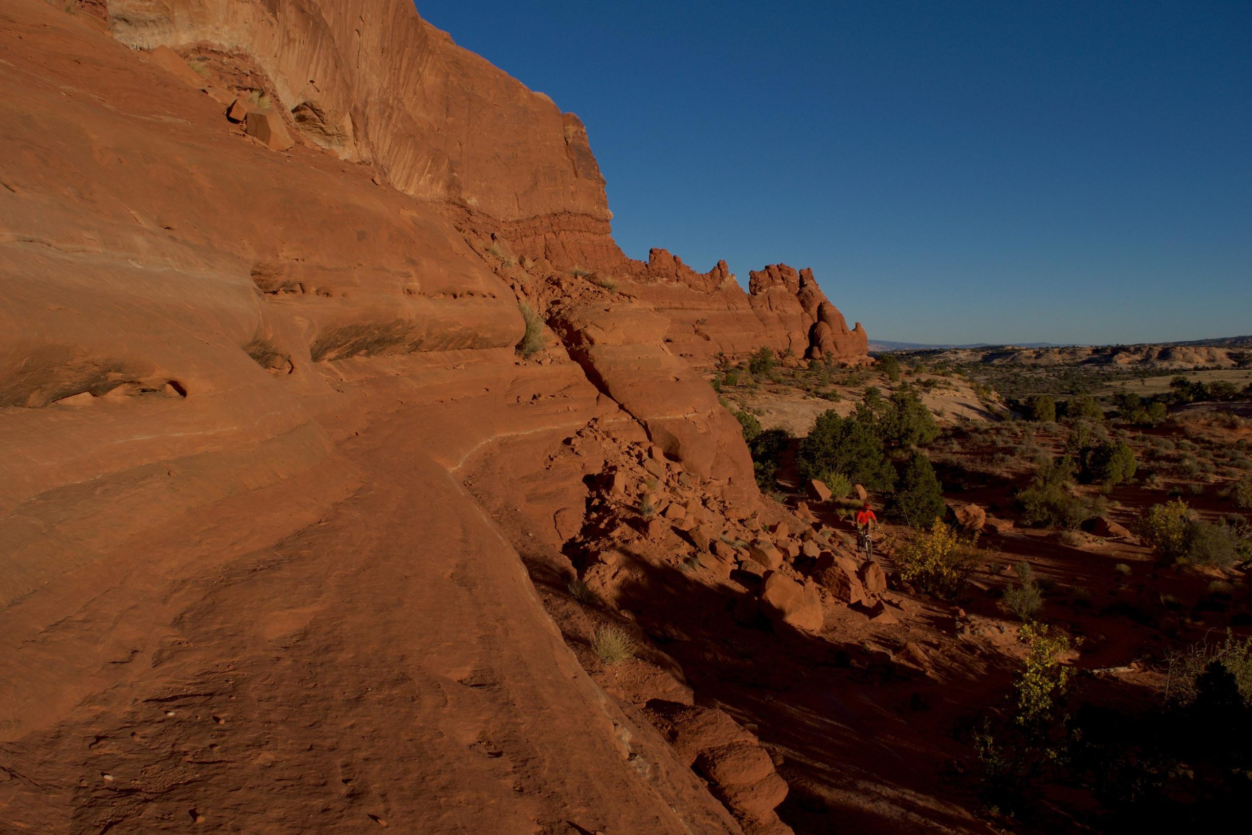 A rugged landscape featuring towering red rock formations under a clear blue sky. The terrain is characterized by smooth, sloping surfaces and scattered vegetation, with a small figure in an orange shirt walking along a narrow path on the lower right. Big Mesa mountain bike trail.