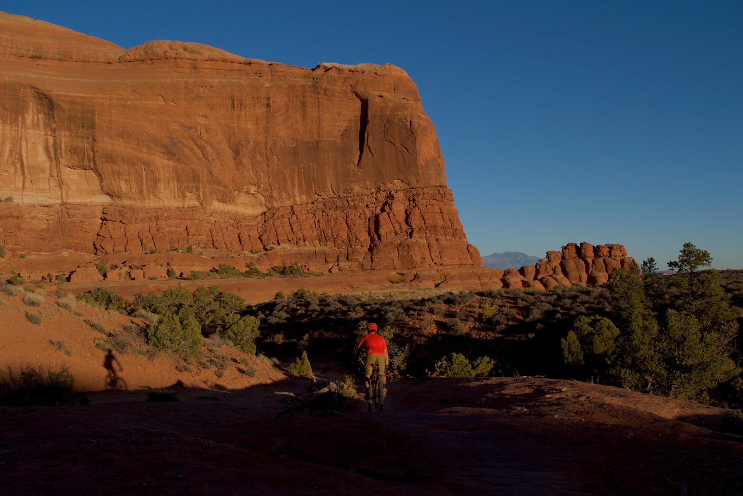 A cyclist riding along a dirt trail in a desert landscape, with towering red rock formations in the background and a clear blue sky above. The cyclist is wearing an orange shirt and helmet, and their shadow is cast on the ground. Sagebrush and sparse vegetation surround the area. Big Mesa mountain bike trail.