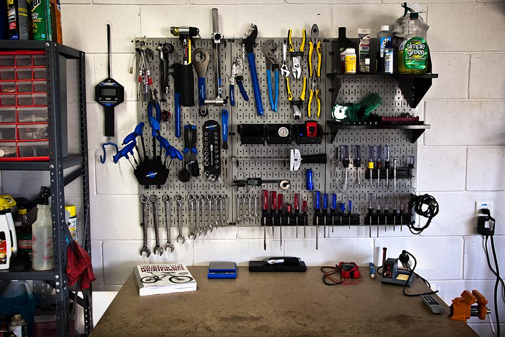 A well-organized garage workspace featuring a pegboard wall with various hand tools, including wrenches, pliers, and screwdrivers. Below the pegboard, a sturdy workbench is visible, with a book on cycling mechanics and several small tools and electronic devices. To the left, a shelving unit contains cleaning supplies and other storage containers. The garage has a clean, functional atmosphere, suitable for DIY projects and repairs.