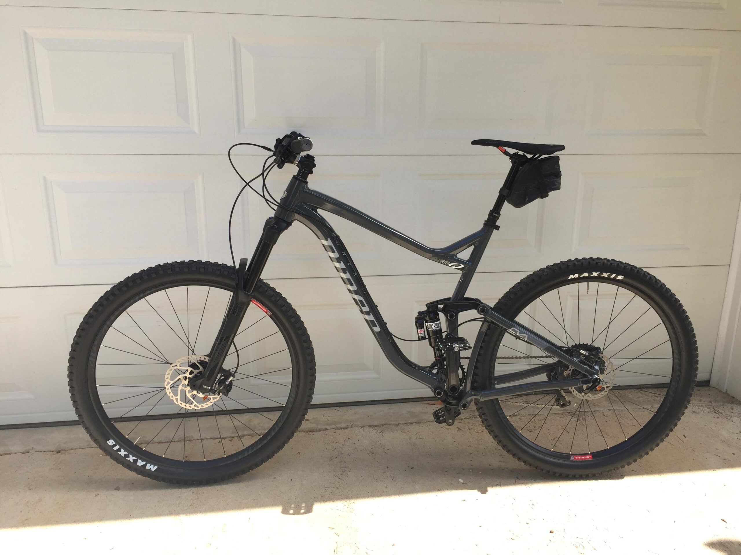 Niner RIP 9: A black mountain bike with wide tires and disc brakes, positioned next to a garage door. The bike features a suspension system, a comfortable seat with a small bag attached, and detailed branding on the frame and tires. The image has a clean background with a concrete surface.