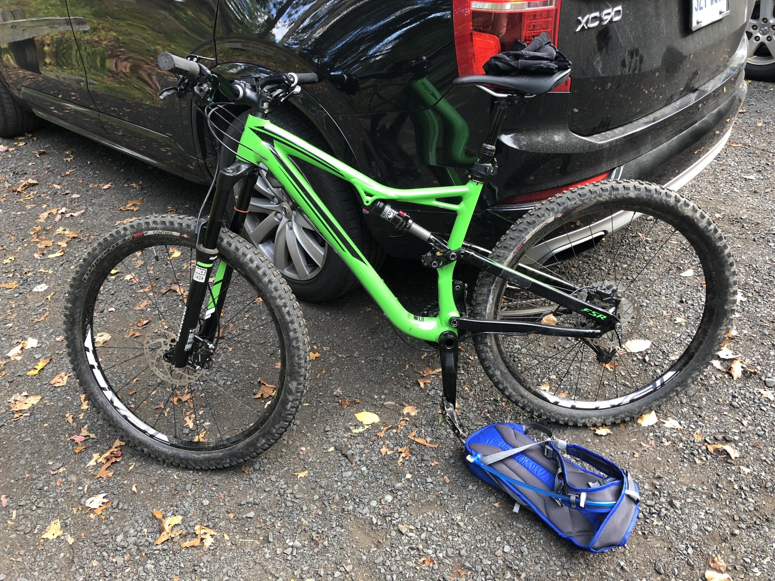 Specialized Stumpjumper FSR: A green and black mountain bike leaning against a black SUV, with a blue bag resting on the gravel surface. Fallen leaves are scattered around the area, indicating a natural outdoor setting.