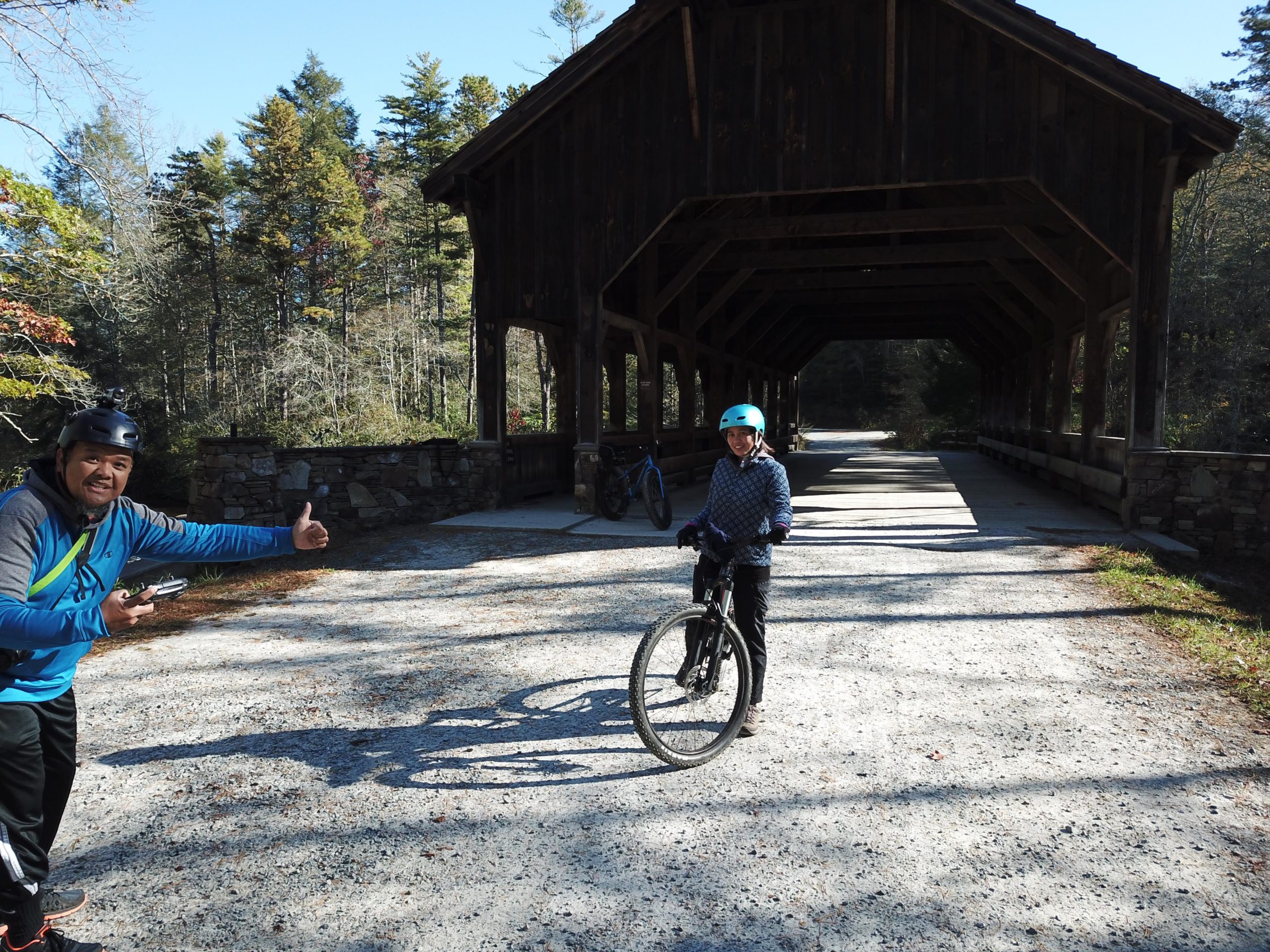 Two cyclists stand near a covered wooden bridge surrounded by trees. One person is giving a thumbs-up while holding a camera, and the other is smiling while standing next to a bicycle. The scene is bright and sunny, highlighting the outdoor recreational atmosphere. DuPont State Recreational Forest mountain bike trail.
