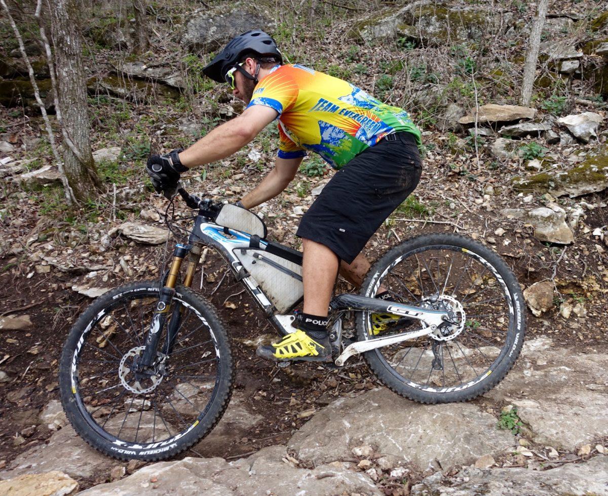 Michelin Wild AM: A cyclist navigating a rocky trail on a mountain bike, wearing a colorful jersey and helmet, focused on maintaining balance while riding through a forested area.