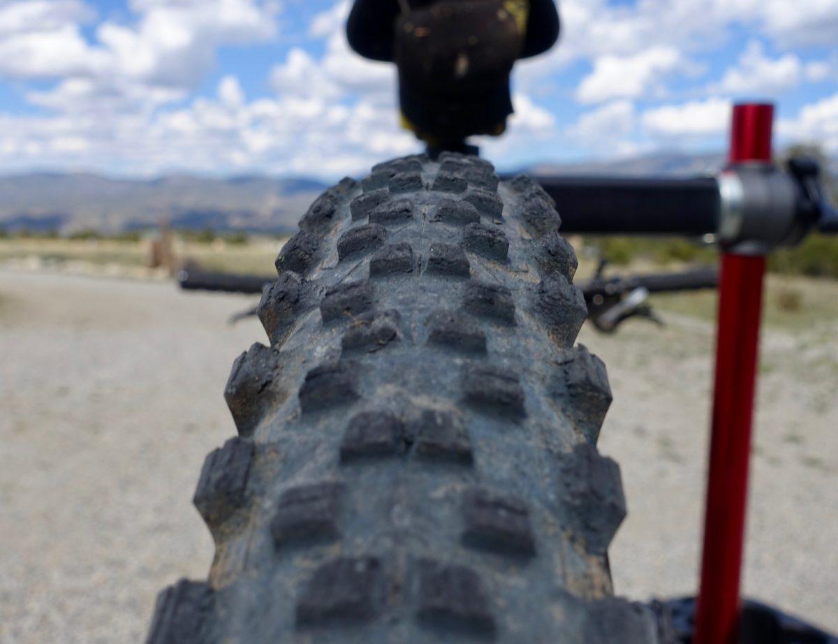 Michelin Wild AM: Close-up view of a mountain bike tire, showcasing its textured tread pattern. In the background, a blurred landscape of hills and a cloudy sky is visible. The bike's handlebar and grip are partially in the frame, indicating the bike is parked or stationary.