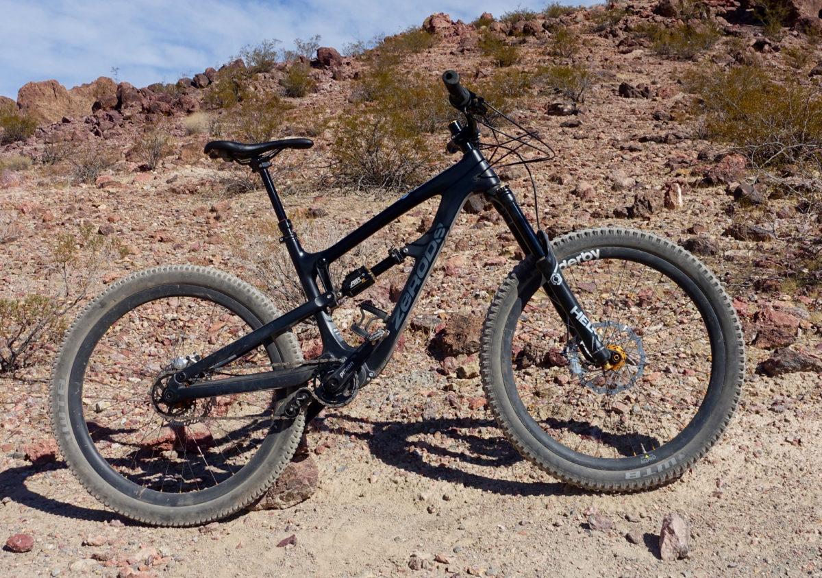 Zerode Taniwha: A black mountain bike resting on a rocky, desert landscape with sparse vegetation and hills in the background. The bike features thick tires and a sturdy frame, showing signs of use on a dirt path.