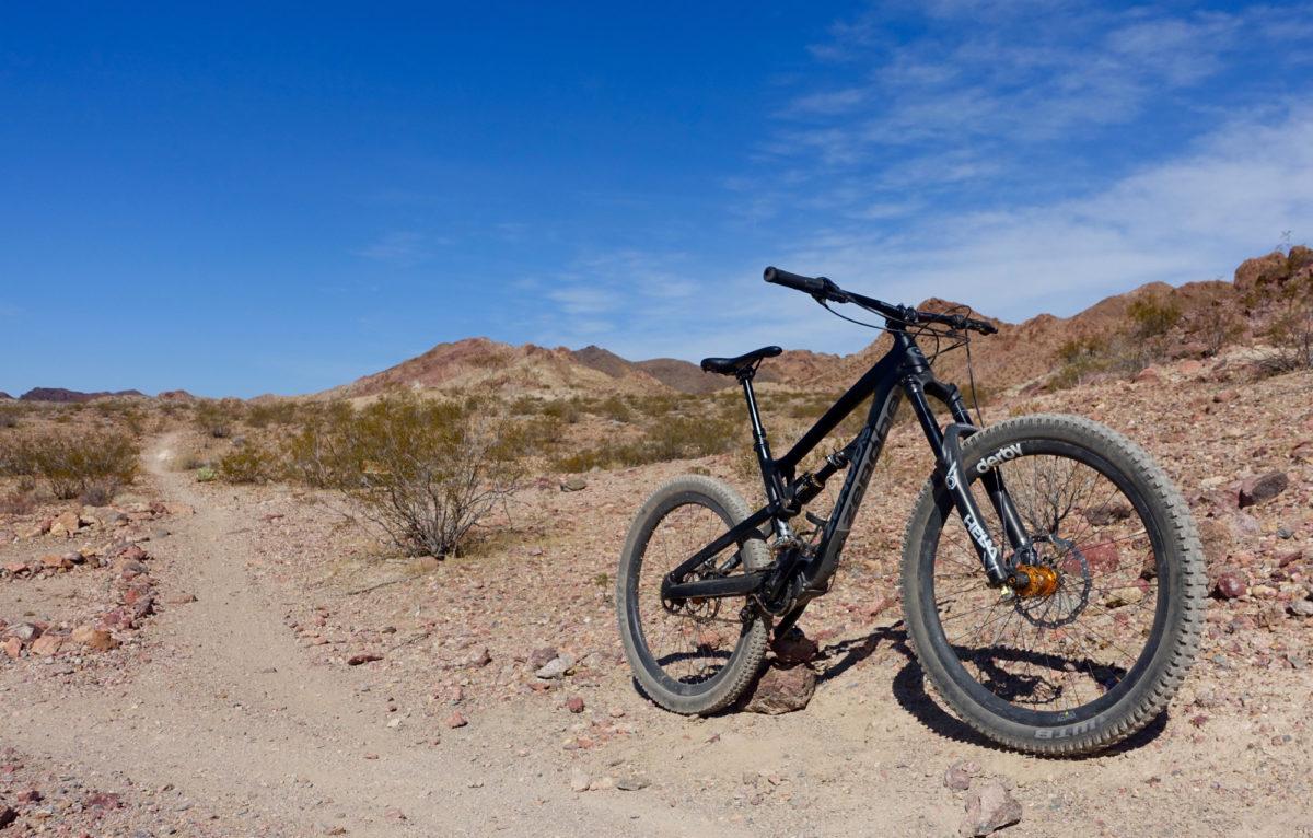 Zerode Taniwha: A black mountain bike is parked on a dirt trail in a rocky desert landscape with sparse vegetation and distant hills under a blue sky with some clouds.