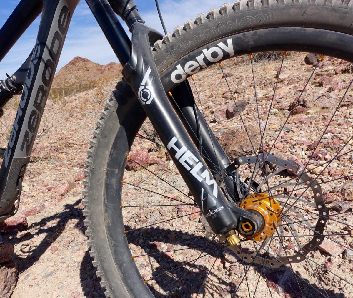 Zerode Taniwha: Close-up view of a mountain bike front fork and wheel on rocky terrain, featuring the brand names "Zerode" and "Helm" on the fork, and a "derby" rim with visible spokes and a disc brake. The background shows rocky hills under a clear blue sky.