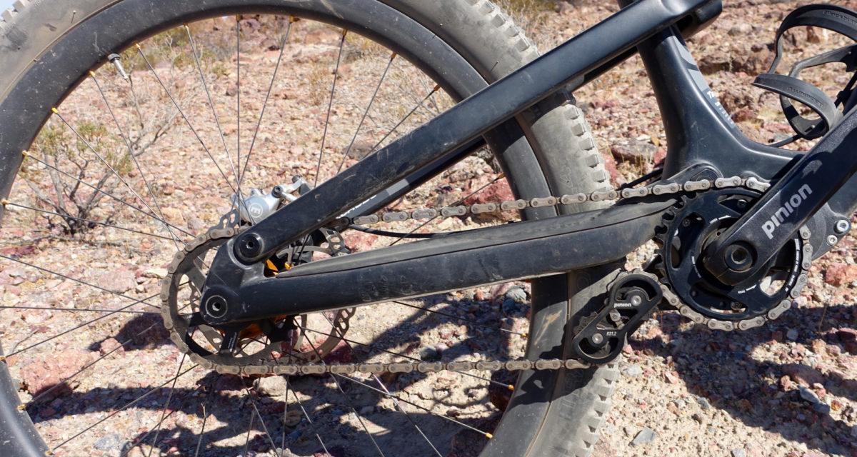 Zerode Taniwha: Close-up view of a mountain bike's drivetrain, featuring a black crankset, chain, and gearing components on a rocky terrain, with a blurred background of sparse vegetation.
