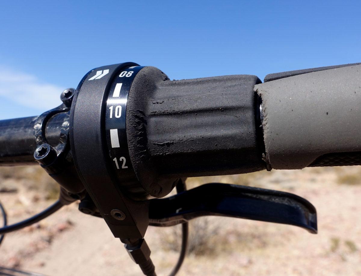 Zerode Taniwha: Close-up of a bicycle handlebar with a gear shifter displaying numbered settings from 08 to 12, alongside textured grips. The background shows a clear blue sky and a blurred landscape of desert terrain.