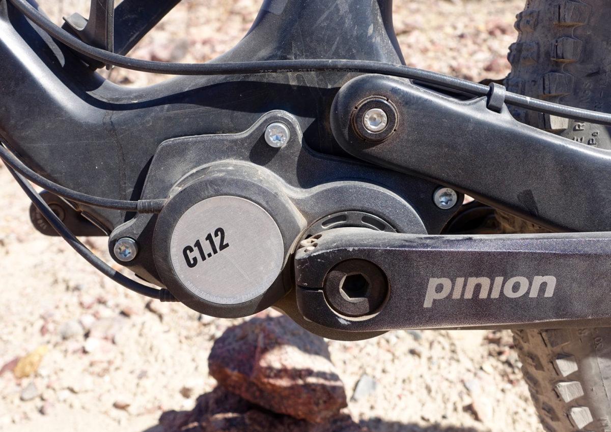 Zerode Taniwha: Close-up view of a bike's crankset and gearbox, showing the Pinion branding and a circular component labeled "C1.12." The background features rocky terrain, suggesting an outdoor setting.