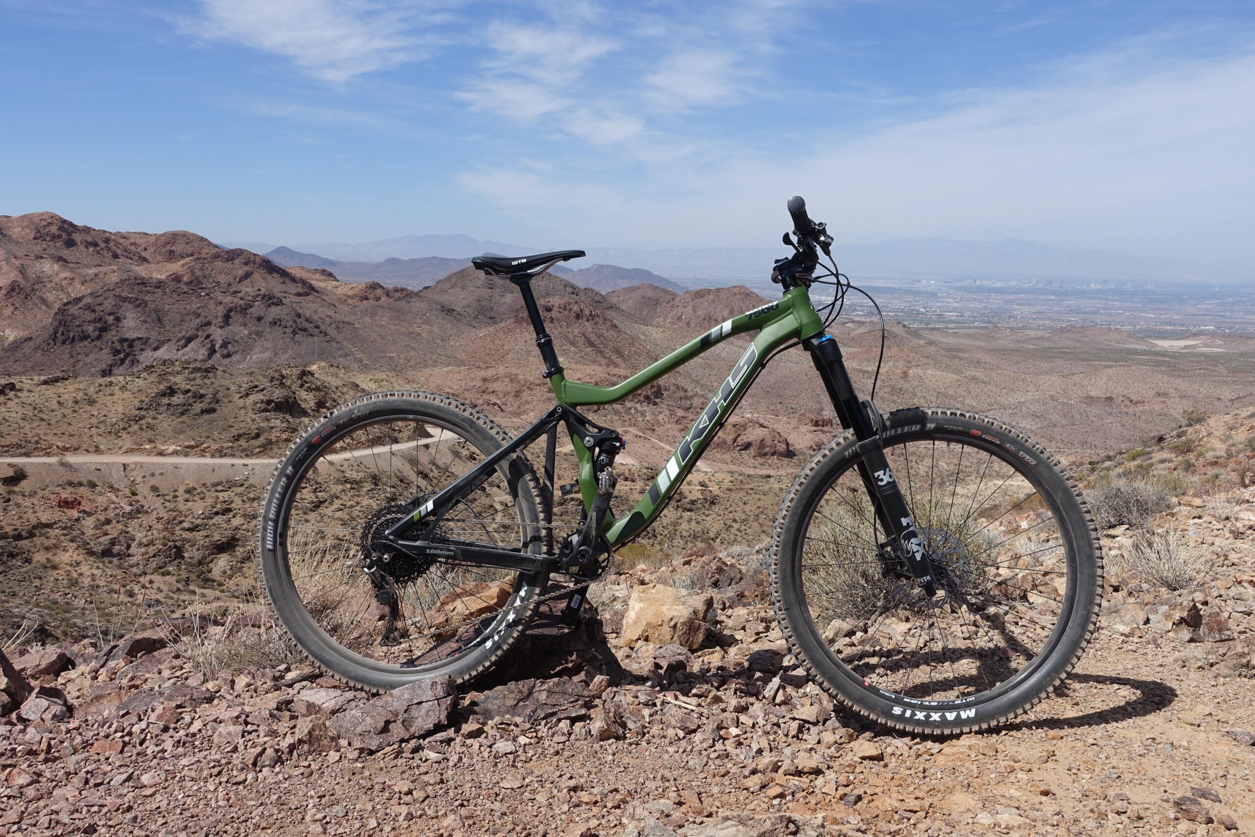 KHS SixFifty 7500: A green mountain bike positioned on rocky terrain with a mountainous landscape in the background and a clear blue sky.