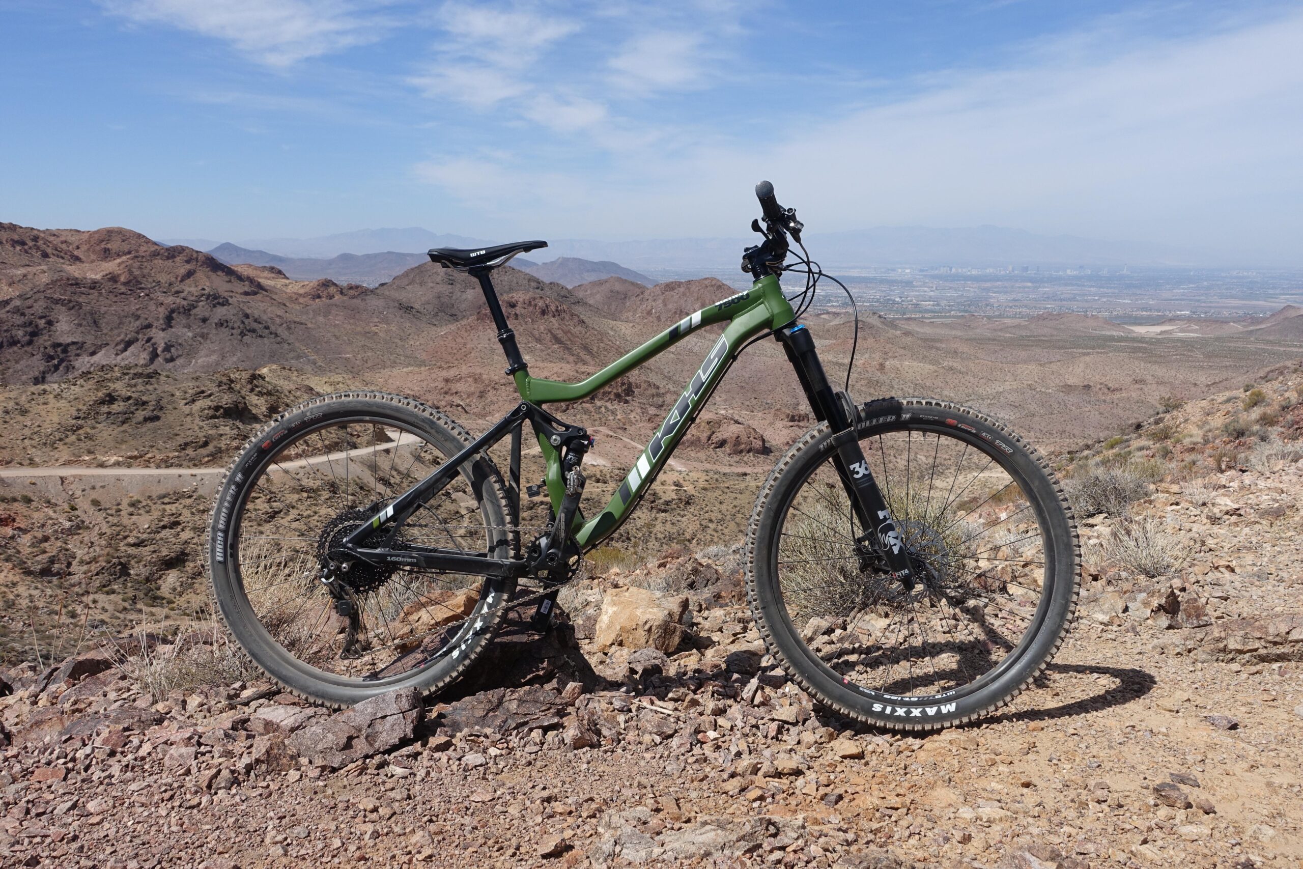 KHS SixFifty 7500: A green and black mountain bike is positioned on rocky terrain with a mountainous landscape in the background. The sky is blue with some clouds, and a distant valley can be seen, indicating a scenic outdoor adventure setting.
