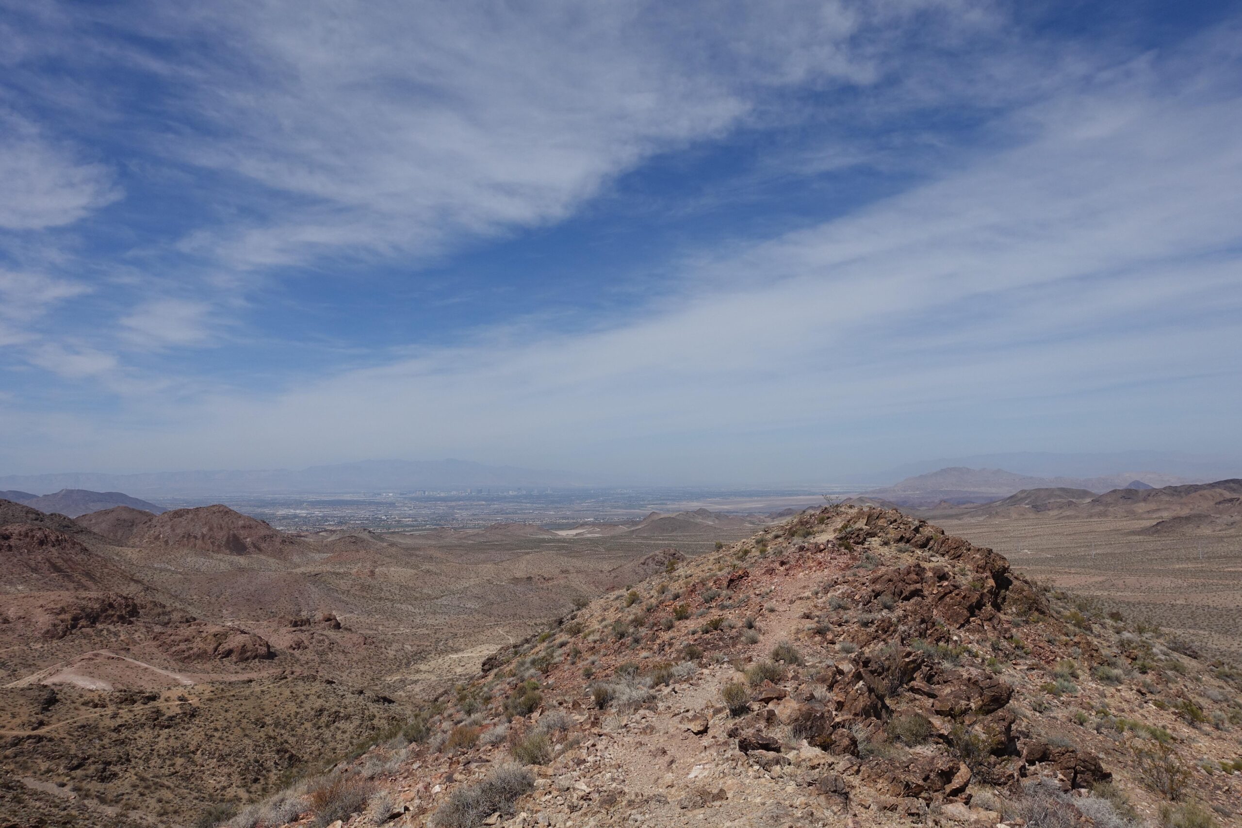 KHS SixFifty 7500: A panoramic view of a rugged desert landscape with rocky hills, sparse vegetation, and distant mountains under a bright blue sky with wispy clouds. The terrain features a mix of brown and tan hues, typical of arid environments, with an expansive valley visible in the distance.
