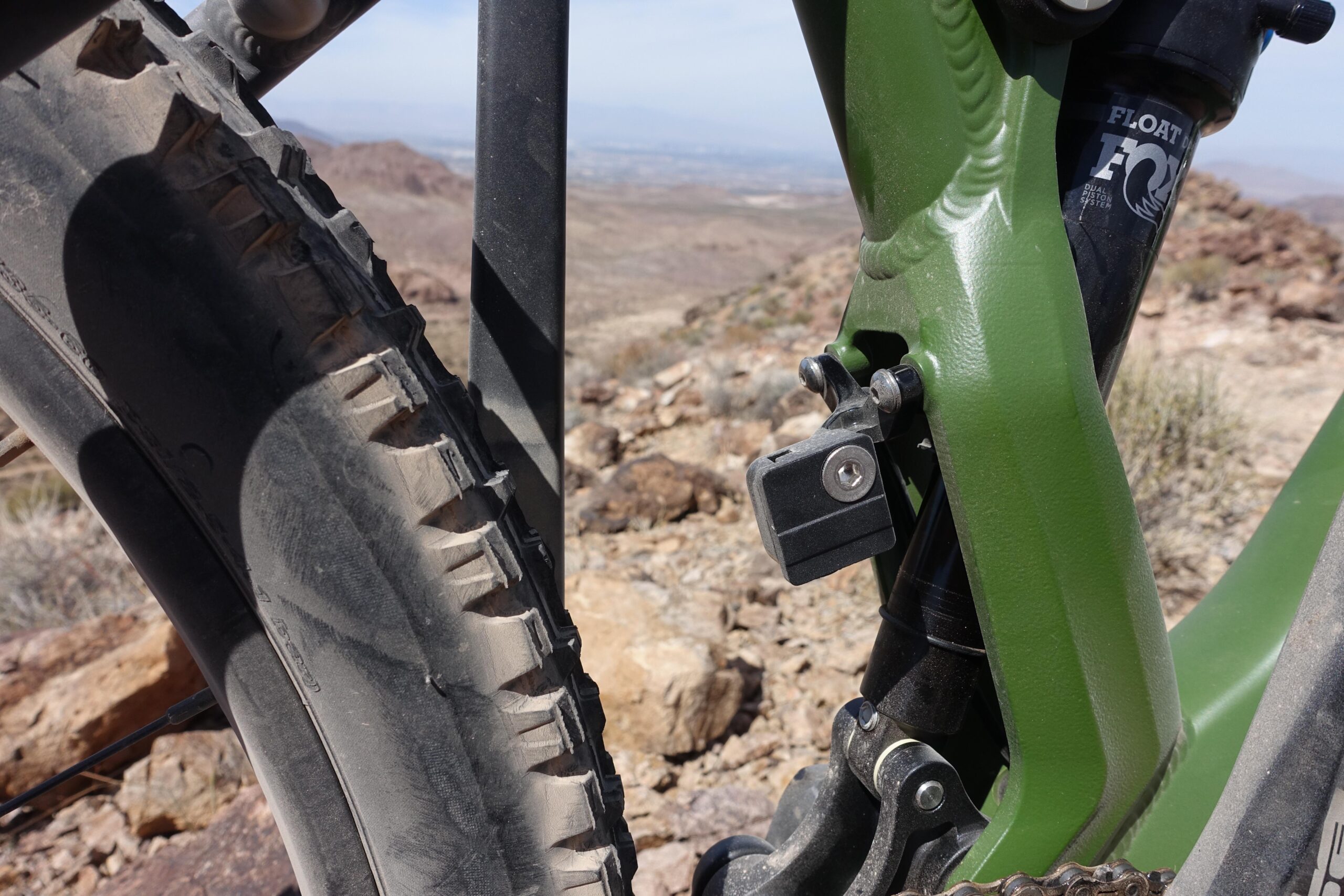 KHS SixFifty 7500: Close-up view of a mountain bike, showcasing the rear wheel and suspension components against a rocky outdoor backdrop. The bike features a green frame, with visible tire tread and a locking mechanism near the suspension. The rugged terrain is evident in the background.