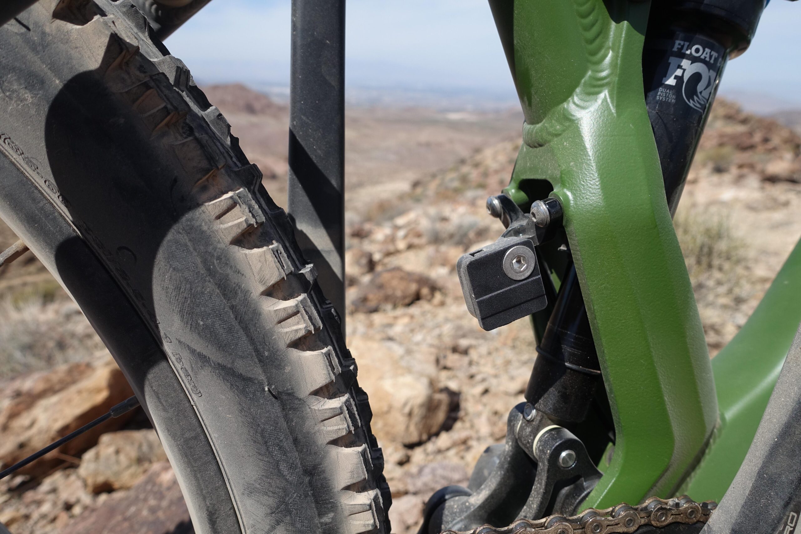 KHS SixFifty 7500: Close-up view of a mountain bike's rear suspension and tire, showcasing a rugged tire tread, a green frame component, and a shock absorber in a rocky outdoor setting.