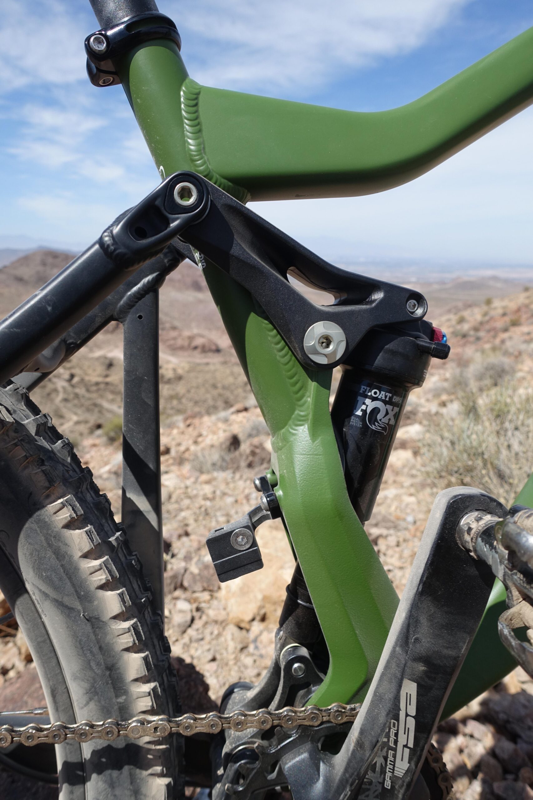 KHS SixFifty 7500: Close-up view of a green mountain bike frame, showcasing the suspension system and components against a natural rocky landscape. The bike's rear suspension and linkages are prominently displayed, with a focus on the black shock absorber labeled "FLOAT DPS FOX." The background features a blurred view of mountains and sky.
