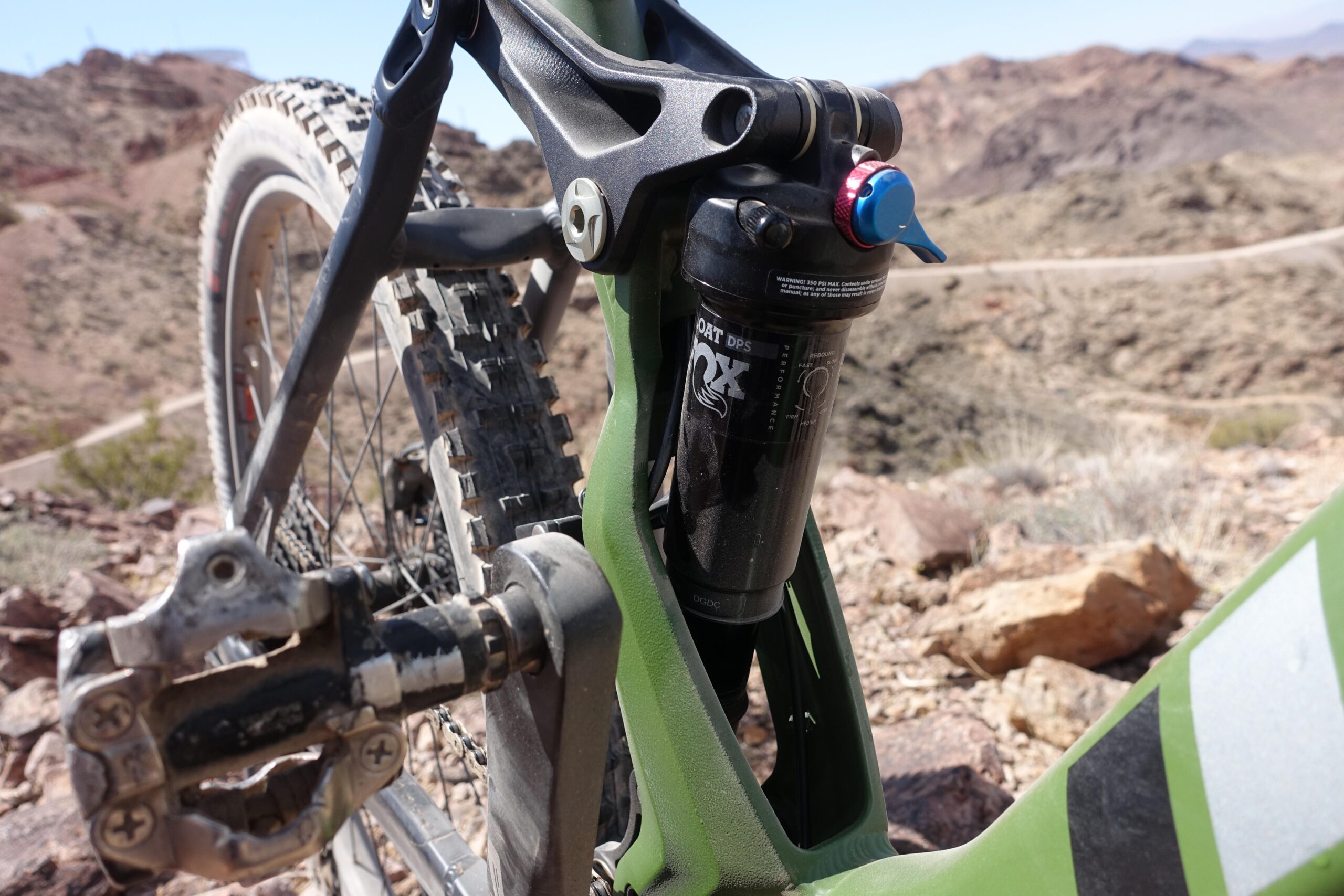 KHS SixFifty 7500: Close-up view of the rear suspension of a mountain bike, showcasing the shock absorber and the pedal. The bike's frame is green, with a portion of the tire visible in the background. The setting includes rocky terrain and mountains in the distance under a clear blue sky.