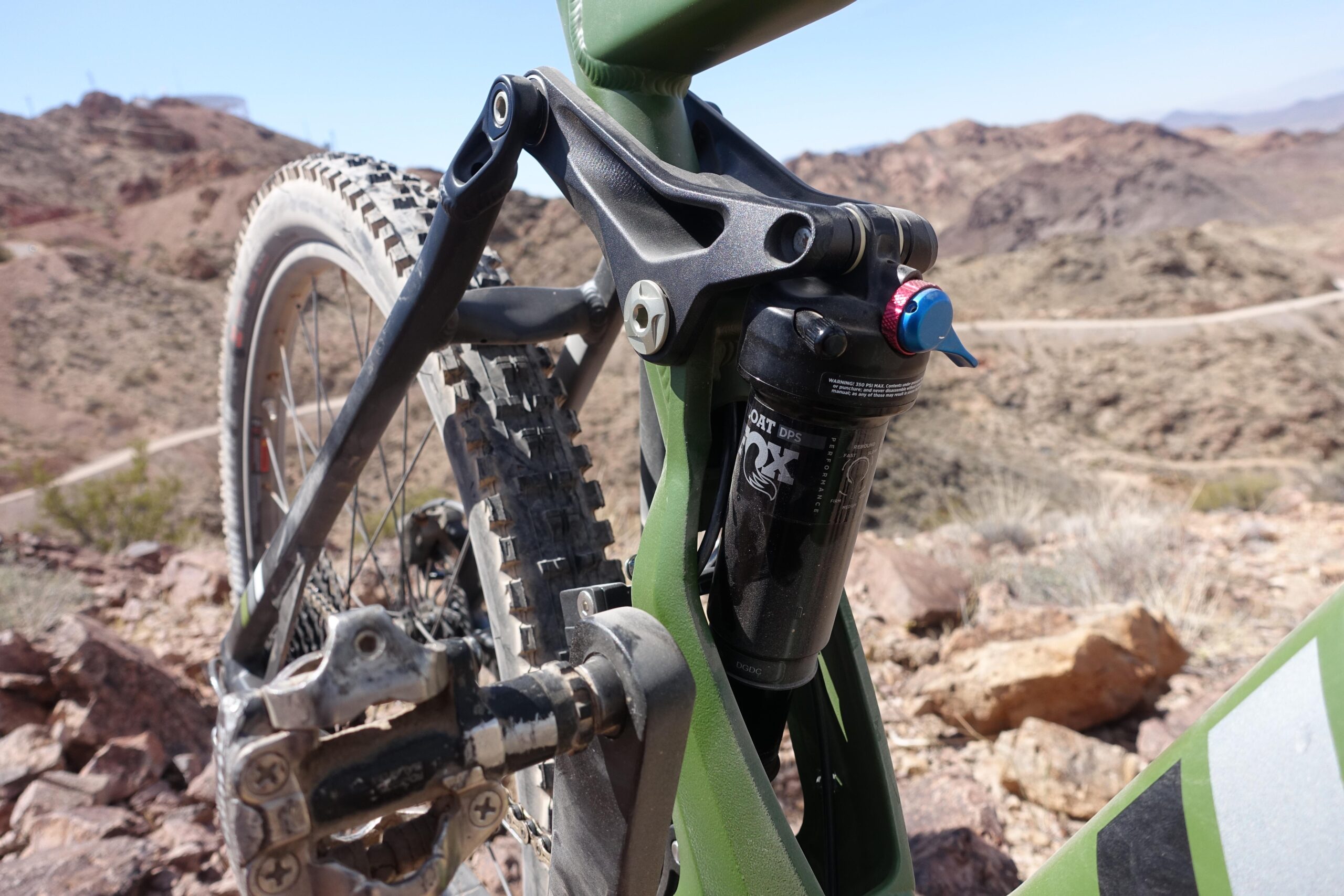 KHS SixFifty 7500: Close-up view of a mountain bike's rear suspension and drivetrain, showcasing the shock absorber and rear triangle against a rugged mountain landscape. The bike frame is primarily green, and the image highlights the detailed components including the chain, pedals, and tire tread.