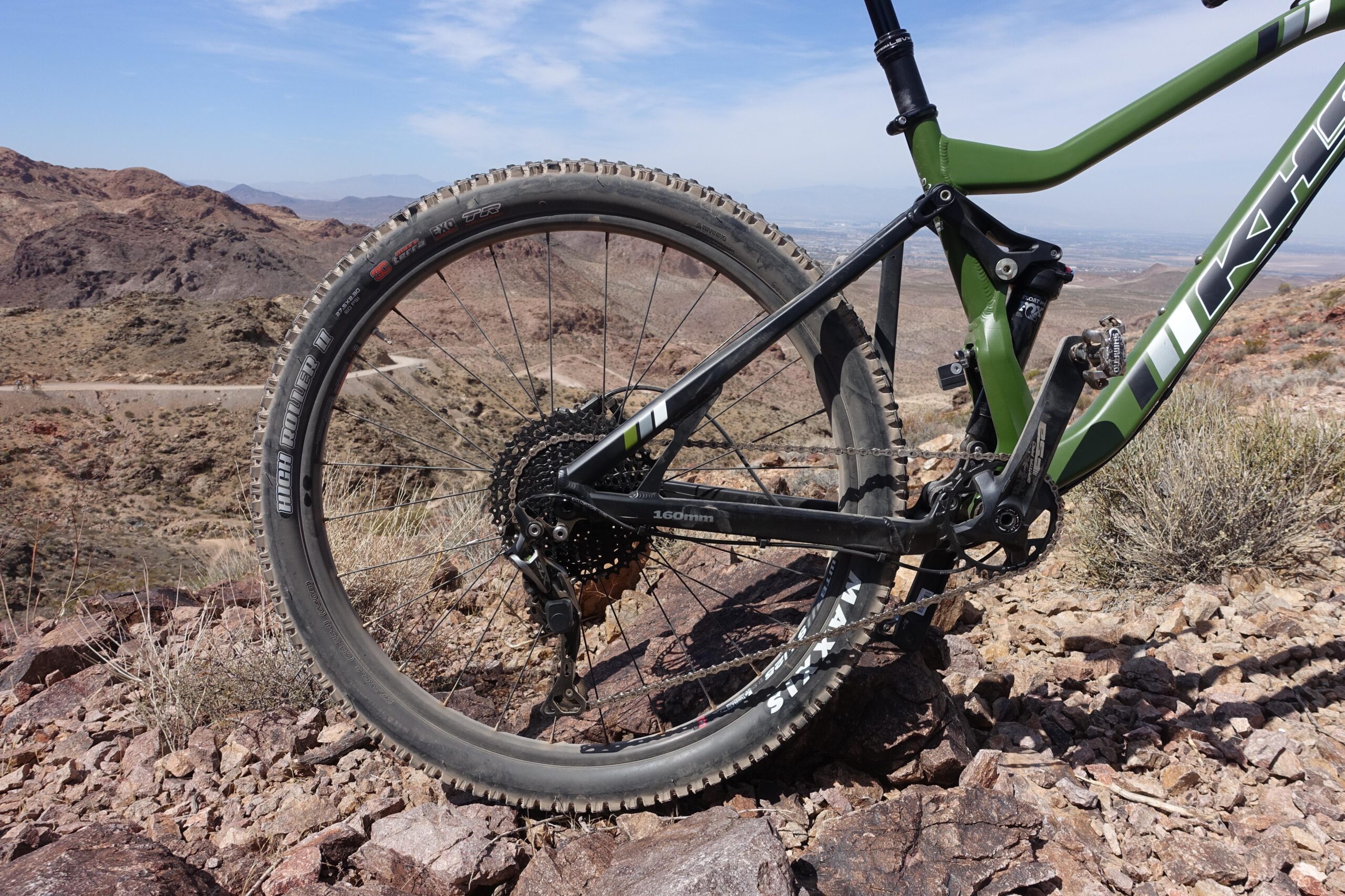 KHS SixFifty 7500: Close-up view of the rear wheel and frame of a mountain bike, set against a rugged desert landscape with rocky terrain and distant mountains under a clear blue sky. The bicycle features a green frame and is equipped with wide tires suitable for off-road riding.