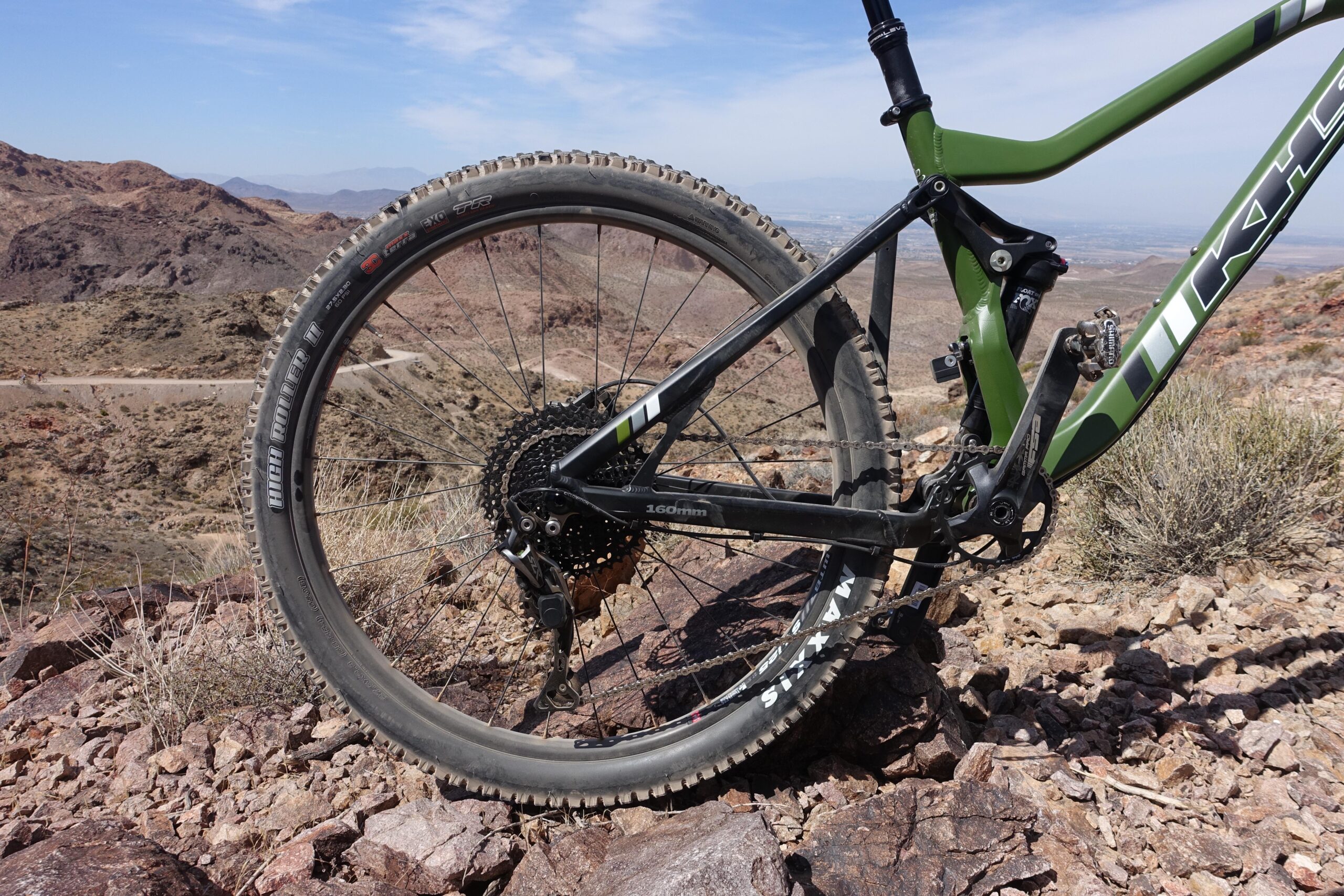 KHS SixFifty 7500: A close-up view of a mountain bike's rear wheel and frame set against a rugged mountainous landscape. The bike features a sleek green and black design, with visible components such as the gears and derailleur. The terrain is rocky and arid, with distant mountains and a clear blue sky in the background.