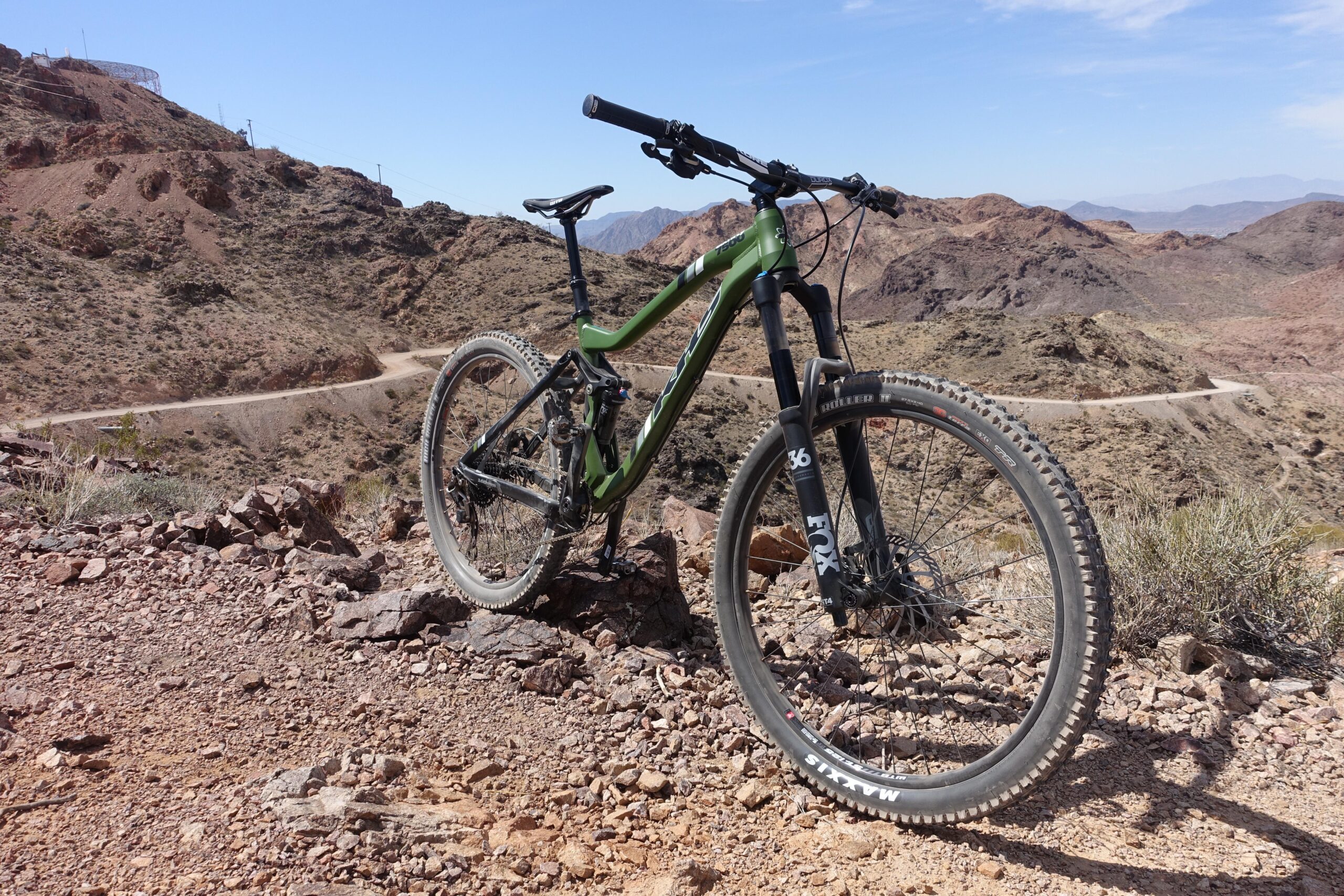 KHS SixFifty 7500: A mountain bike positioned on rocky terrain, with a winding dirt road visible in the background. The setting features rugged hills and clear blue skies, suggesting an outdoor adventure in a hilly or mountainous area.