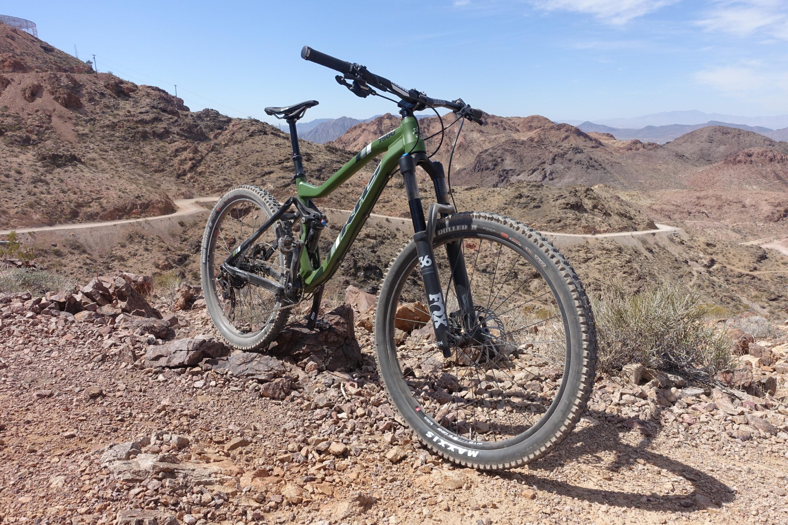 KHS SixFifty 7500: A green mountain bike leaning against a rocky outcrop on a mountain trail, surrounded by arid terrain and distant hills under a clear blue sky.