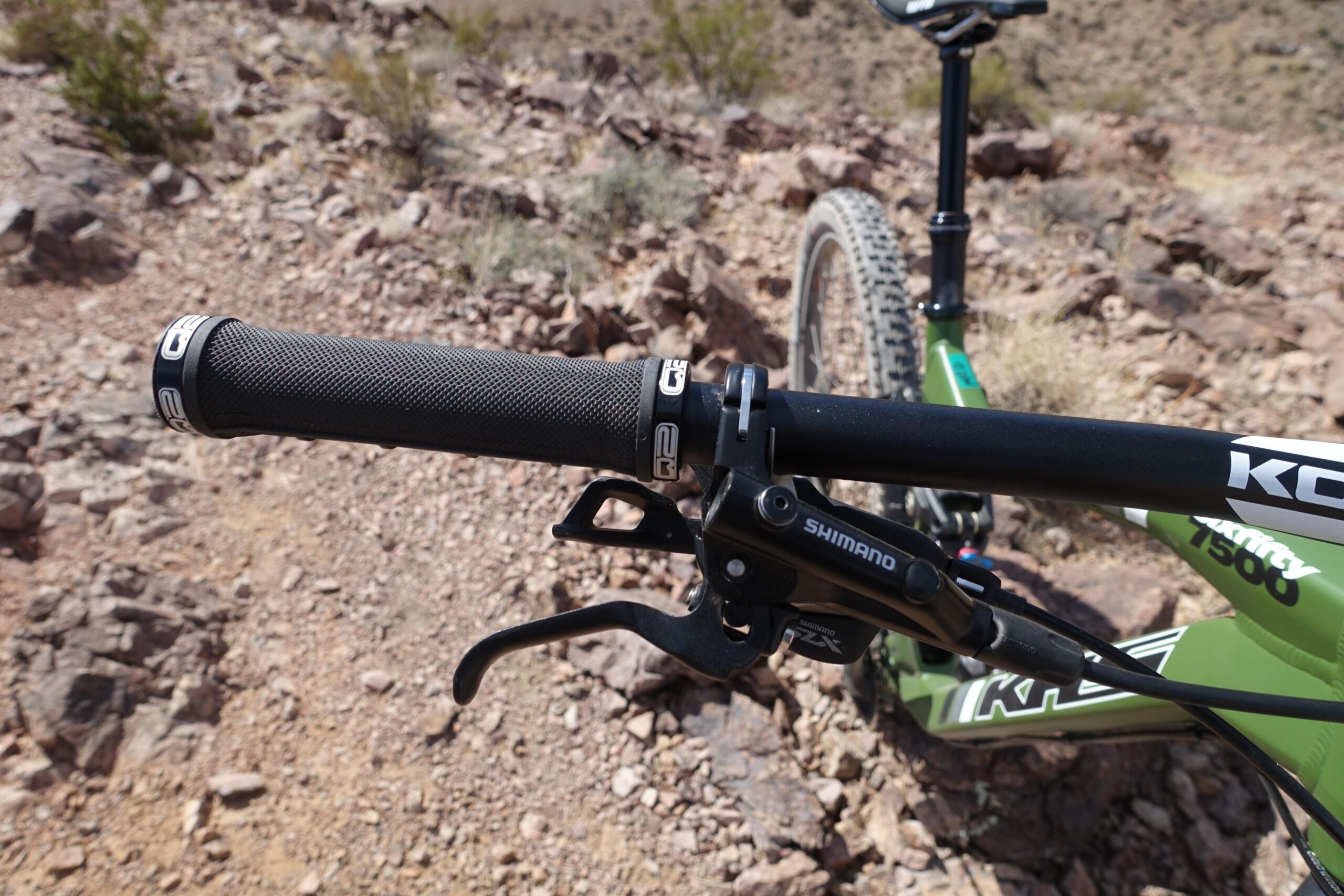 KHS SixFifty 7500: Close-up view of a mountain bike handlebar, featuring a textured black grip, Shimano brake lever, and gear shifter. The background shows a rocky terrain, indicating an outdoor biking environment.