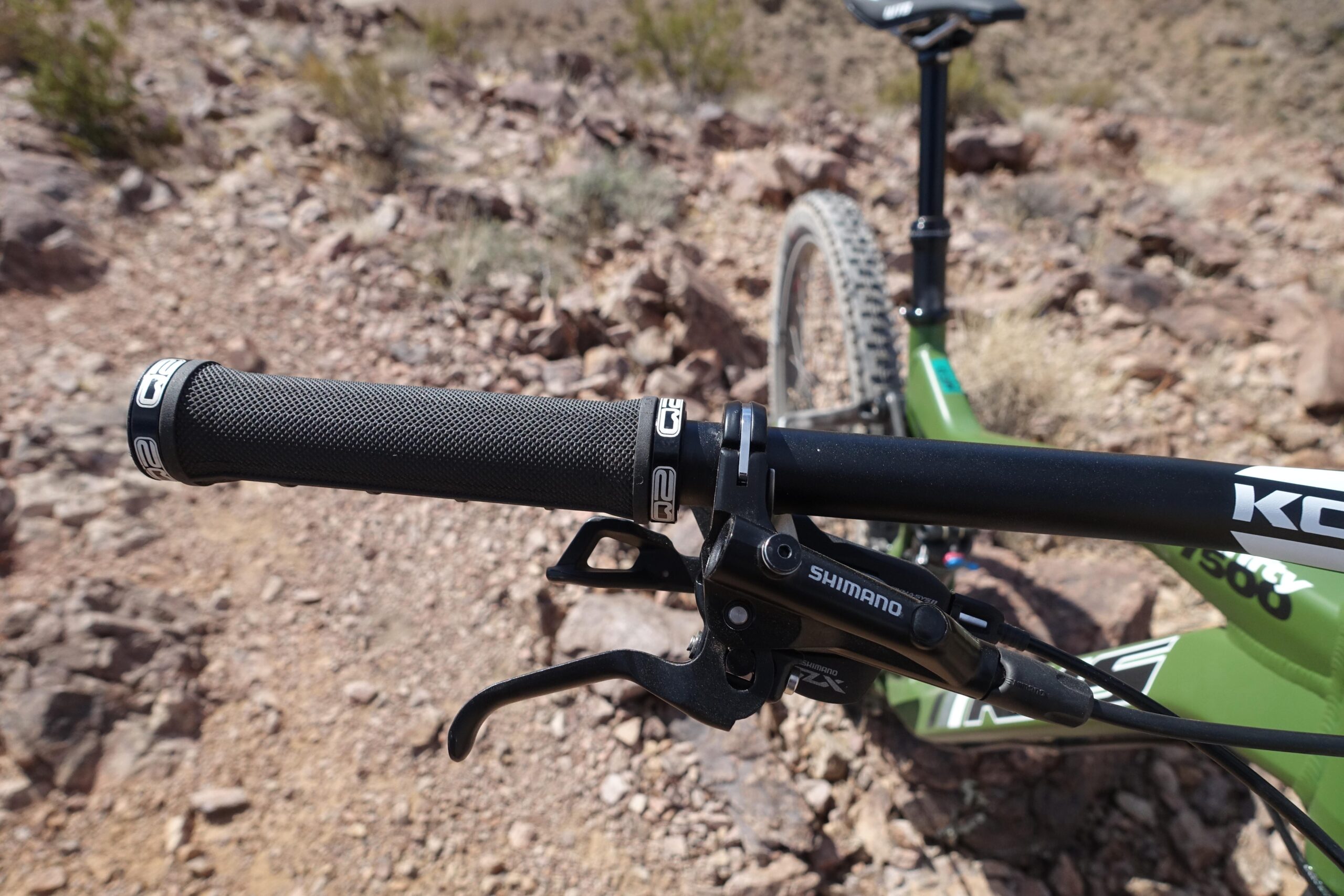 KHS SixFifty 7500: Close-up view of a mountain bike handlebar featuring a textured black grip and Shimano brake lever. The background shows a rocky trail, indicating a rugged outdoor setting.