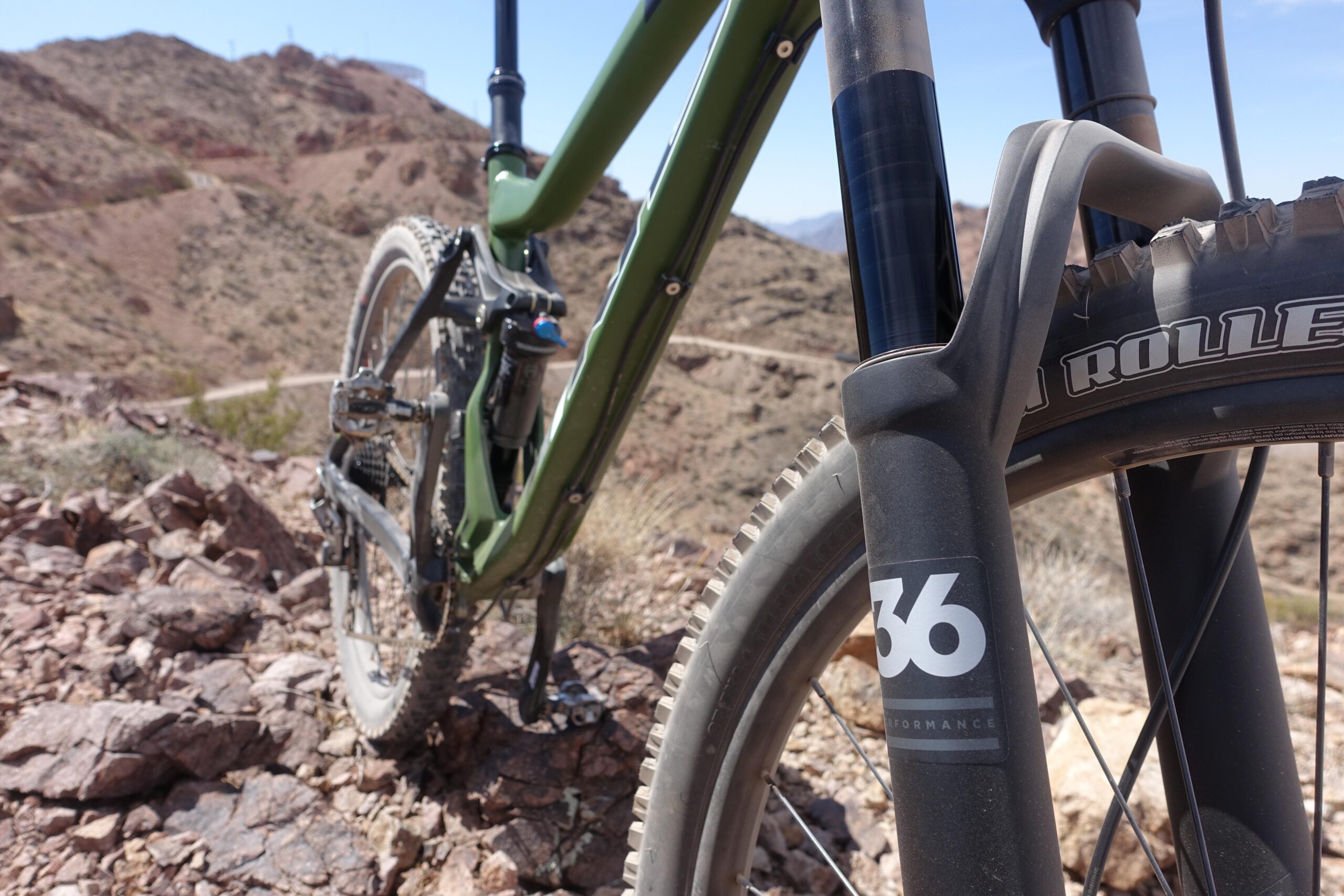 KHS SixFifty 7500: Close-up view of a mountain bike on rocky terrain, showcasing the front fork and tire. The bike has a green frame, and the tire is labeled "Roller." The background features a rugged landscape with hills and a blue sky.