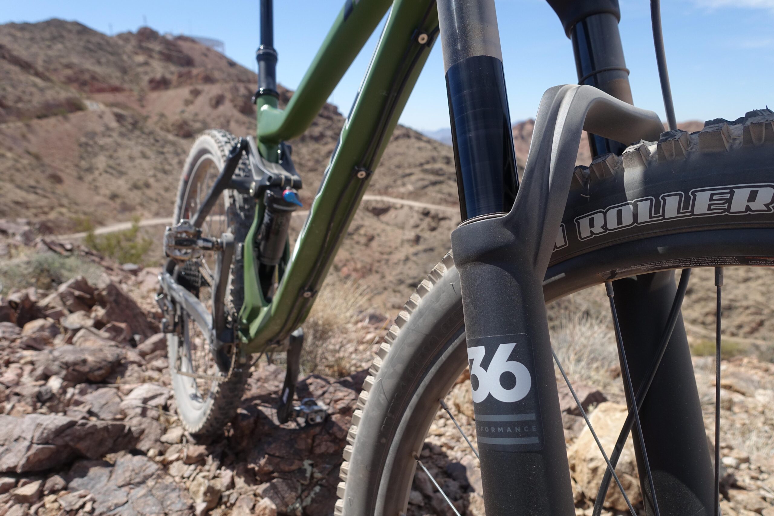 KHS SixFifty 7500: A close-up shot of a mountain bike on rocky terrain, showcasing the front fork and tire. The bike features a green frame and a black fork with a visible "36" performance label. Background includes a rugged landscape with hills and a dirt path.