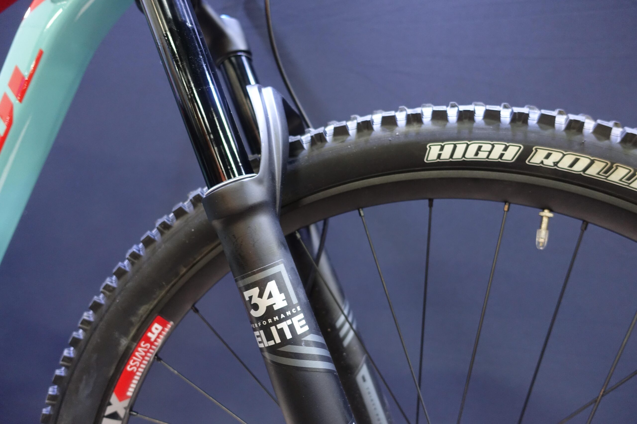 Patrol 591: Close-up of a mountain bike's front suspension fork and tire, featuring a black fork labeled "34 Performance Elite" and a tire with "High Roller" branding, set against a dark blue background.