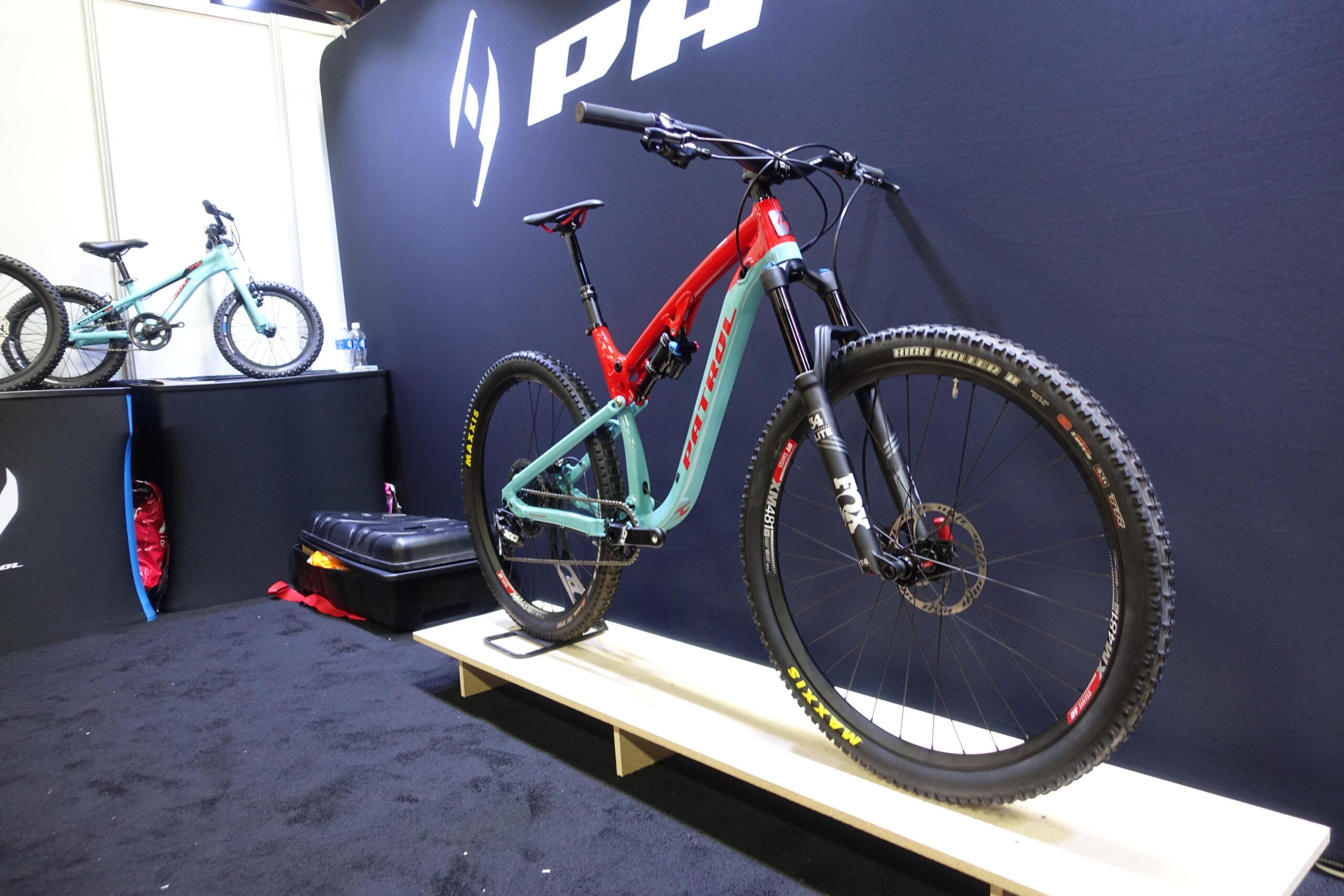 Patrol 591: A red and turquoise mountain bike displayed on a wooden platform against a black backdrop featuring a logo. In the background, a smaller bike and a black storage box are visible, suggesting a bike showroom or exhibition setting.