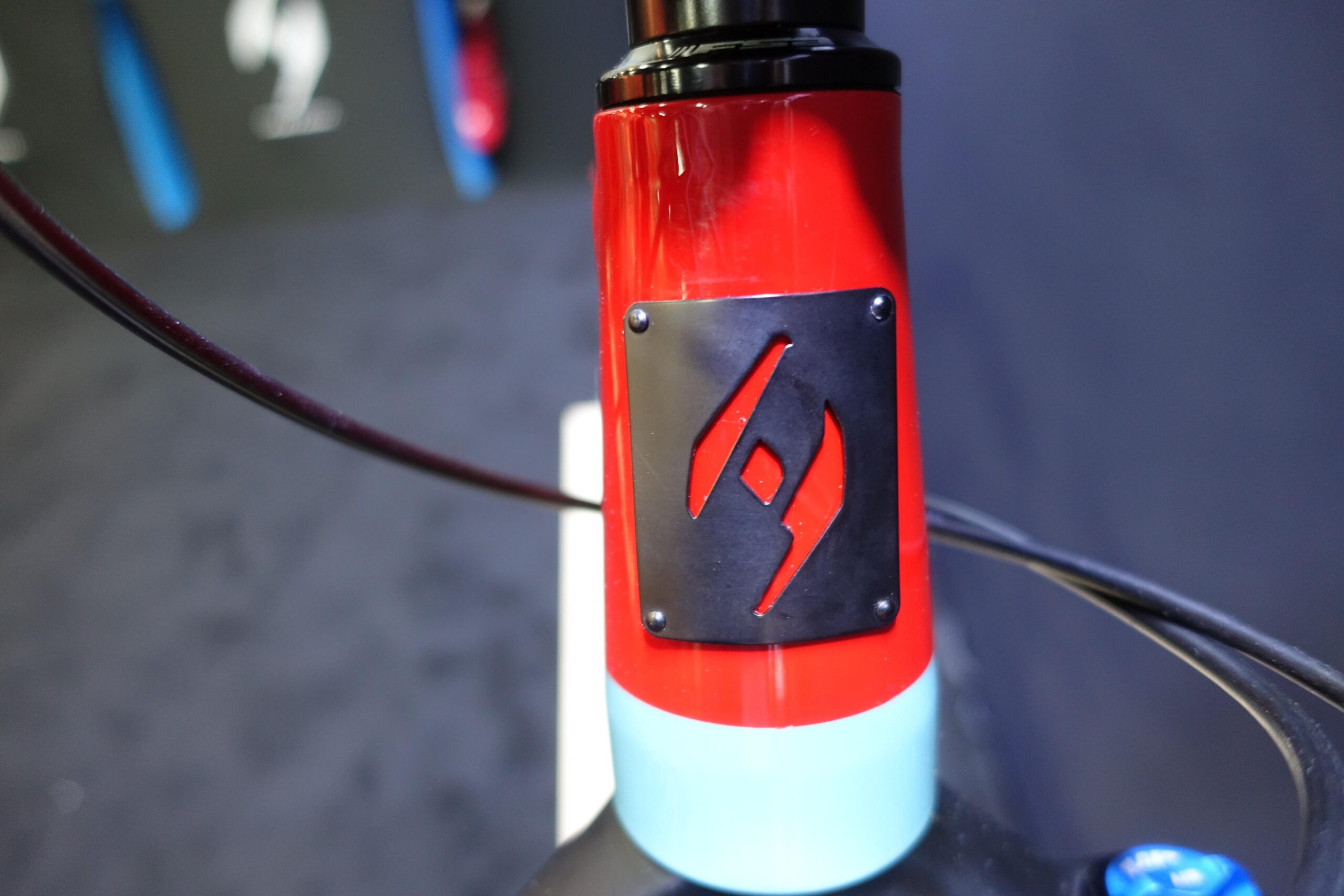 Patrol 591: Close-up image of the top tube of a bicycle, featuring a glossy red finish with a blue section near the bottom. A metallic black plate with a stylized logo is prominently displayed on the front of the frame. The background is softly blurred, emphasizing the bicycle's design details.